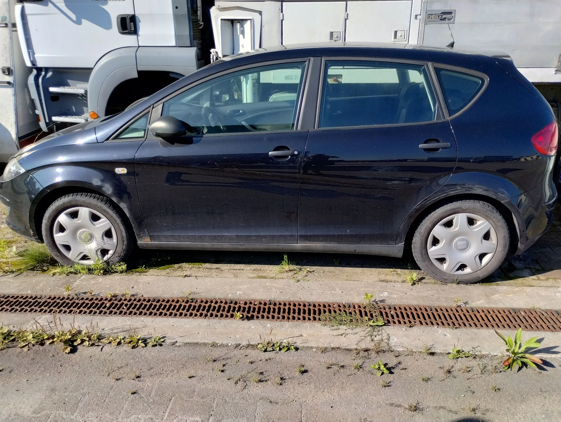 Profil ciemnoniebieskiego samochodu osobowego typu hatchback zaparkowanego na betonowym podłożu obok białej ciężarówki. Pod samochodem widoczna jest metalowa kratka ściekowa z trawą wyrastającą w szczelinach.