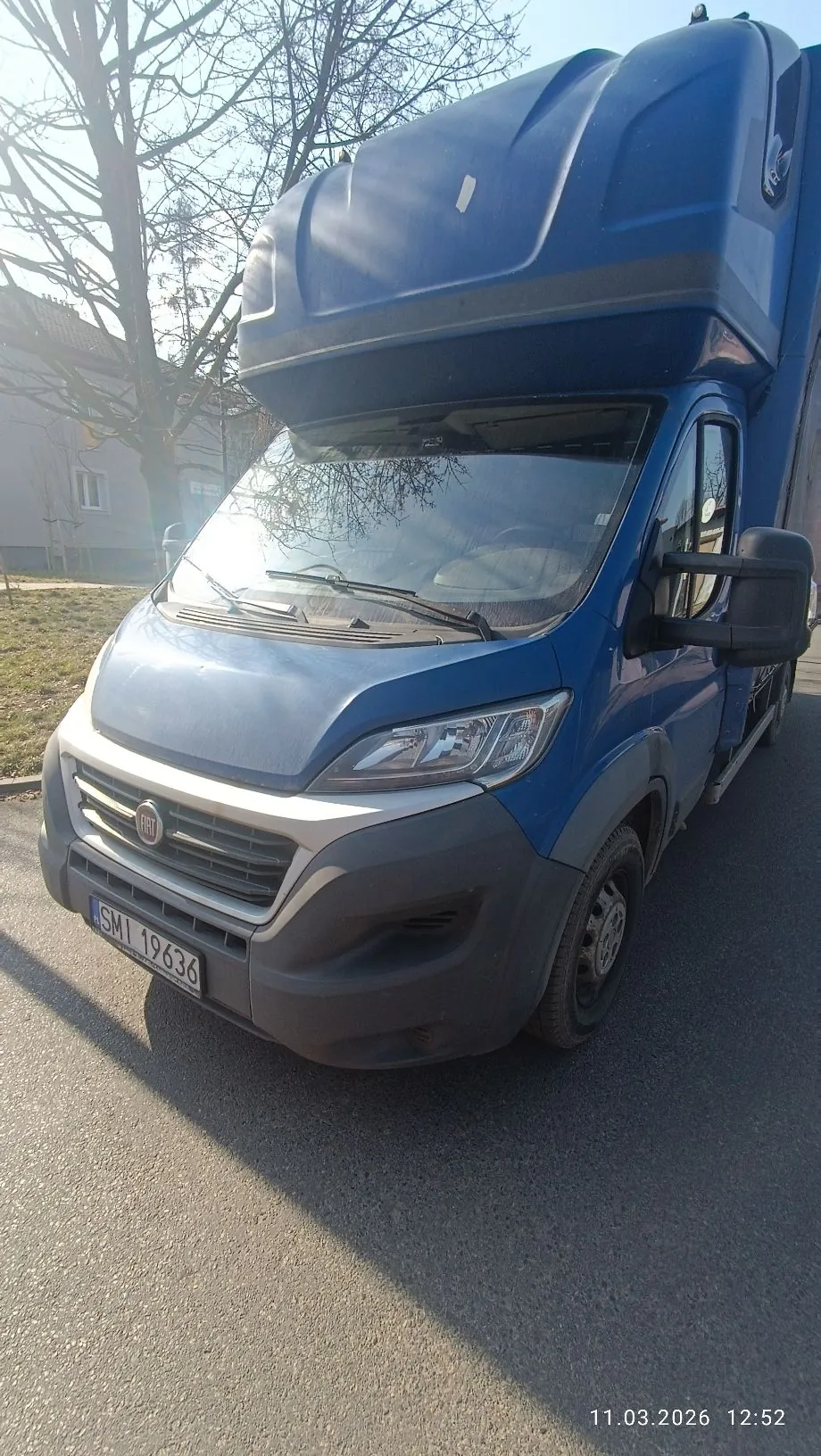 Niebieski samochód dostawczy marki Fiat z wysoką nadbudową nad kabiną kierowcy. Pojazd posiada szary zderzak i tablicę rejestracyjną o numerze SMI 19636. Zdjęcie wykonane na zewnątrz w słoneczny dzień, w tle widoczne są bezlistne drzewa i fragment budynku. W prawym dolnym rogu znajduje się data i godzina: 11.03.2026 12:52.
