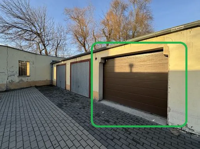 Zdjęcie przedstawia rzęd garaży z zamkniętymi bramami. Po lewej stronie widoczny jest budynek z małym oknem z kratą, a na pierwszym planie brukowana powierzchnia. W tle rosną bezlistne drzewa i niebo. Trzy garaże mają różne kolory bram: szarą, szarą z czerwoną listwą oraz brązową z zielonym prostokątnym zaznaczeniem. Oświetlenie wskazuje na poranek lub późne popołudnie.