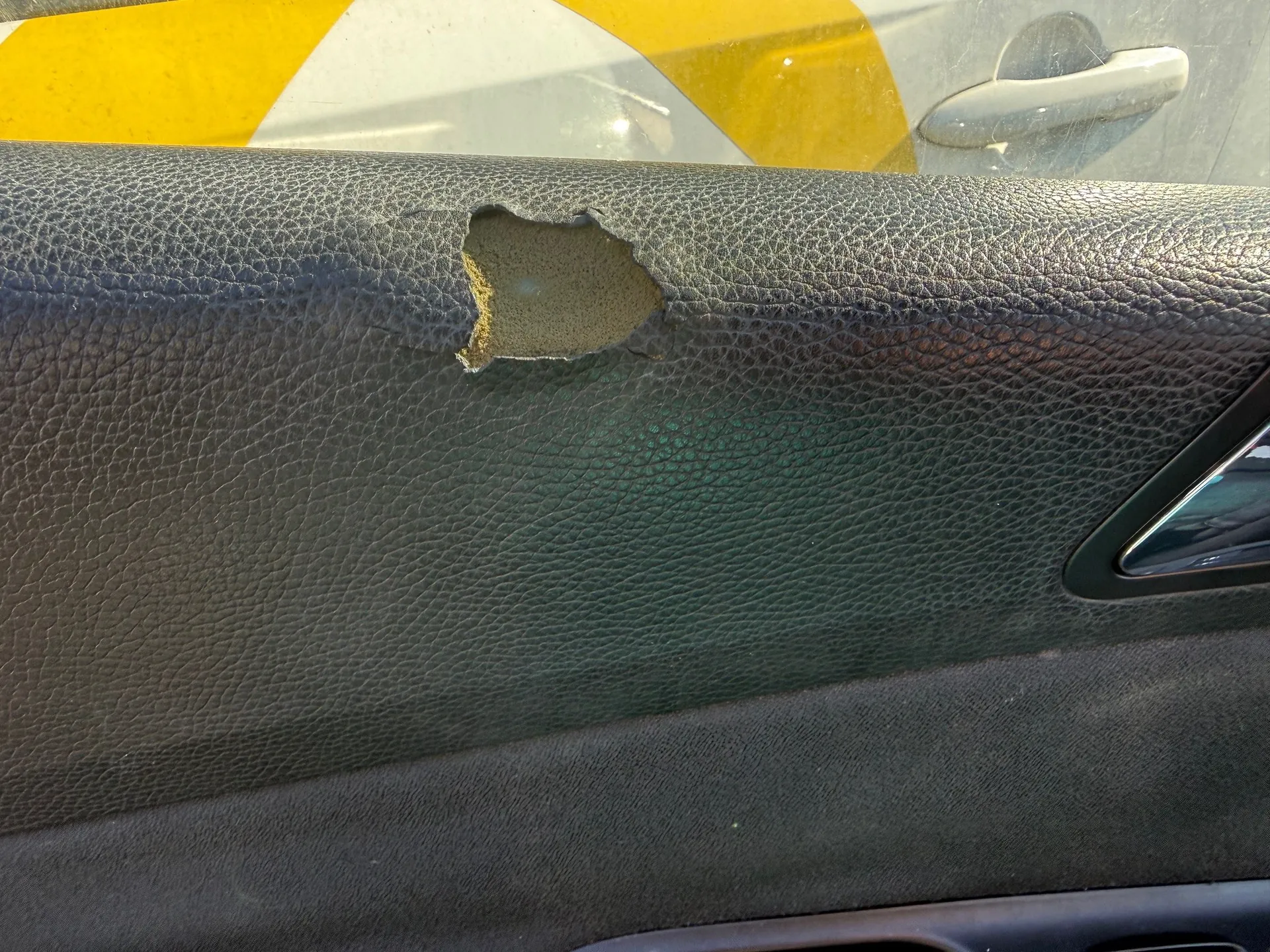Wnętrze drzwi samochodu z uszkodzonym czarnym skórzanym panelem, w którym znajduje się dziura odsłaniająca żółtawą piankę. W tle przez okno widoczne są żółto-białe pasy oraz srebrna klamka drzwi.