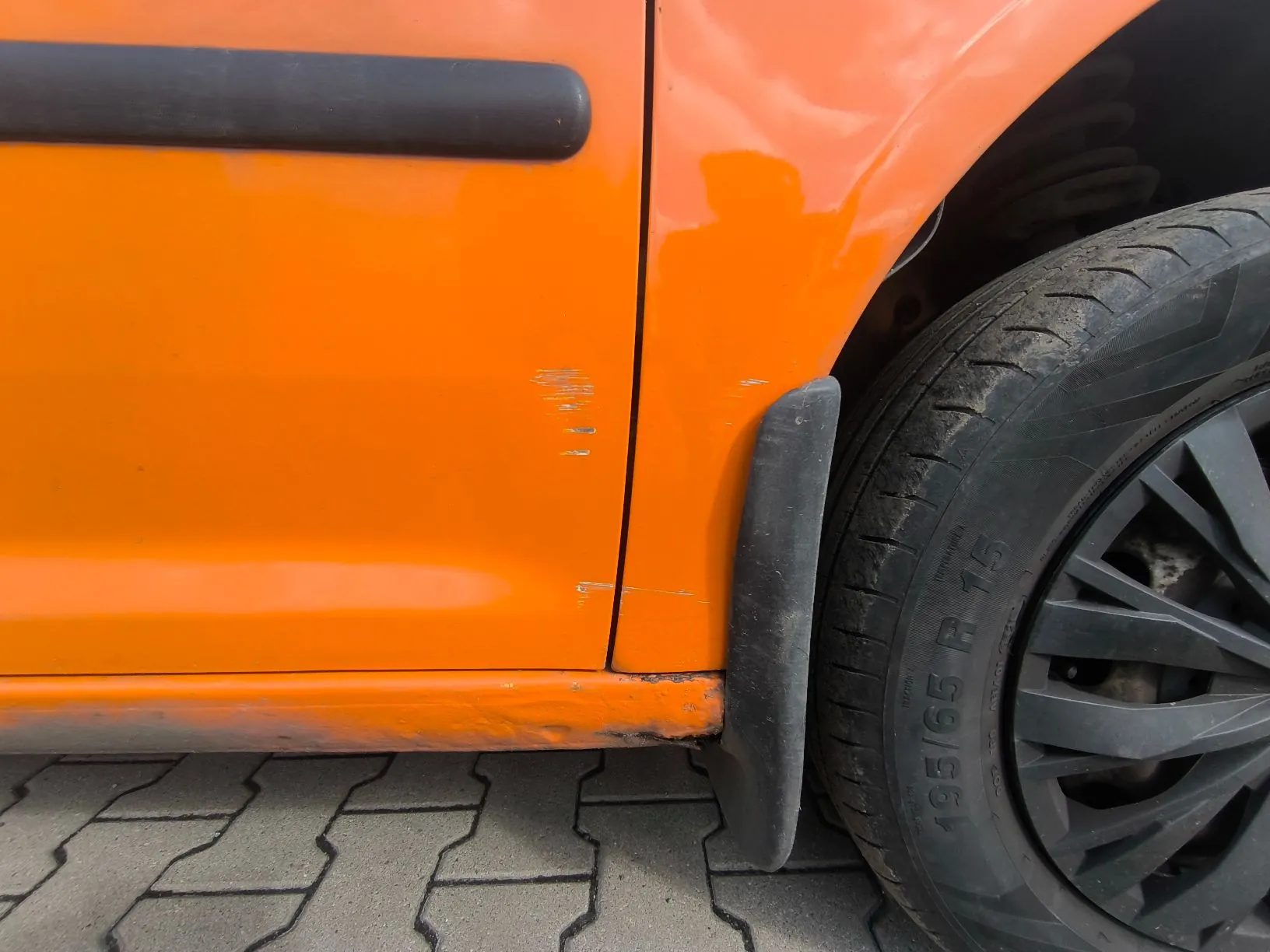 Zbliżenie na pomarańczowy lakier samochodu w okolicy przedniego błotnika i drzwi. Na karoserii widoczne są liczne zarysowania i otarcia, szczególnie w miejscu łączenia drzwi z błotnikiem oraz na dolnej krawędzi progu. Po prawej stronie widoczny jest fragment czarnej opony z napisem '195/65 R 15' oraz czarna felga. Samochód stoi na szarej kostce brukowej.