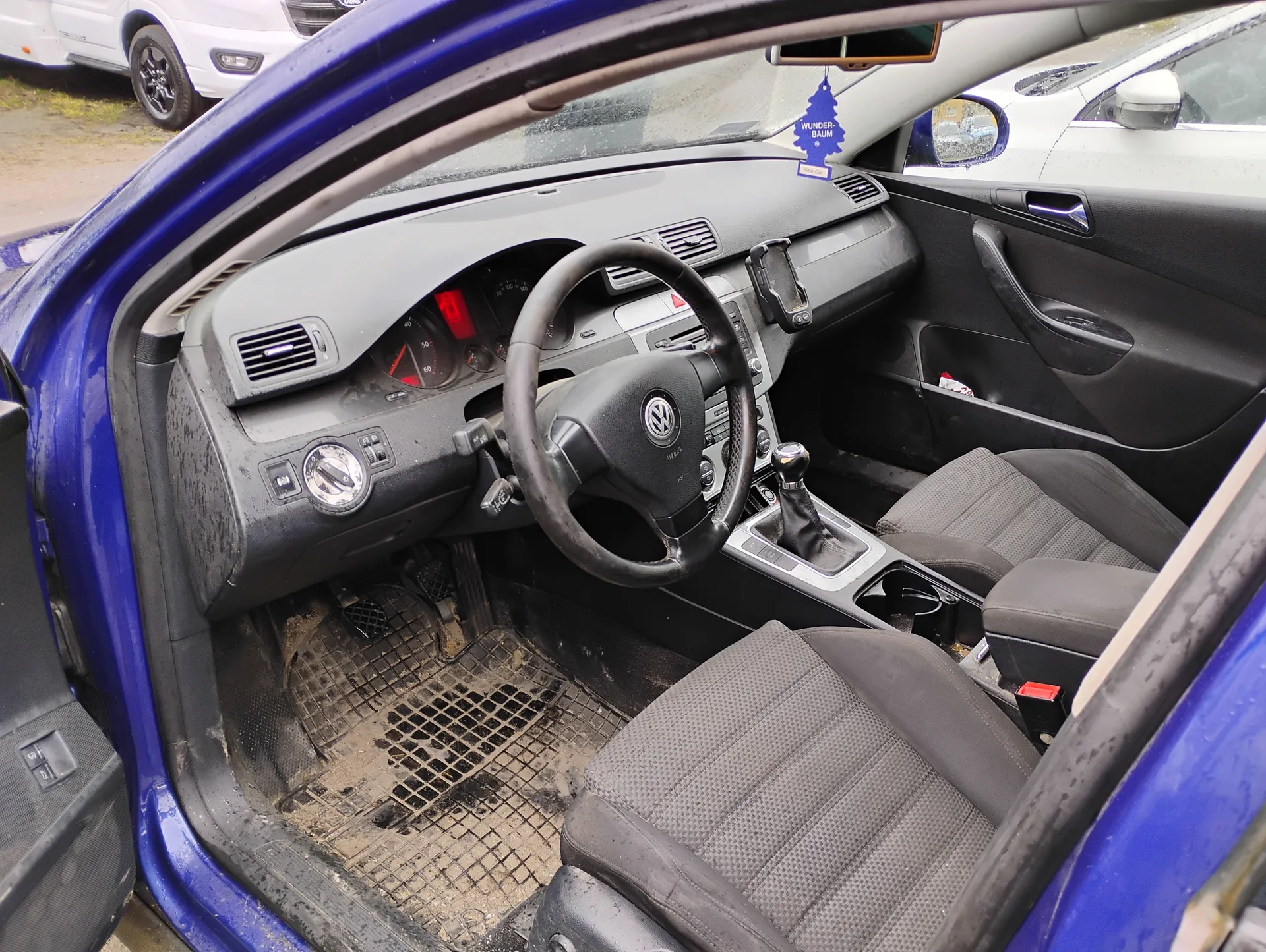 Wnętrze niebieskiego samochodu marki Volkswagen z otwartą drzwią kierowcy. Widoczna kierownica z logo VW, deska rozdzielcza z czerwonymi wskaźnikami, przyciski klimatyzacji, dźwignia zmiany biegów oraz brudne dywaniki podłogowe. Na lusterku wstecznym wiszący niebieski odświeżacz powietrza z napisem "WUNDER-BAUM". Poza samochodem widoczne są inne zaparkowane pojazdy.