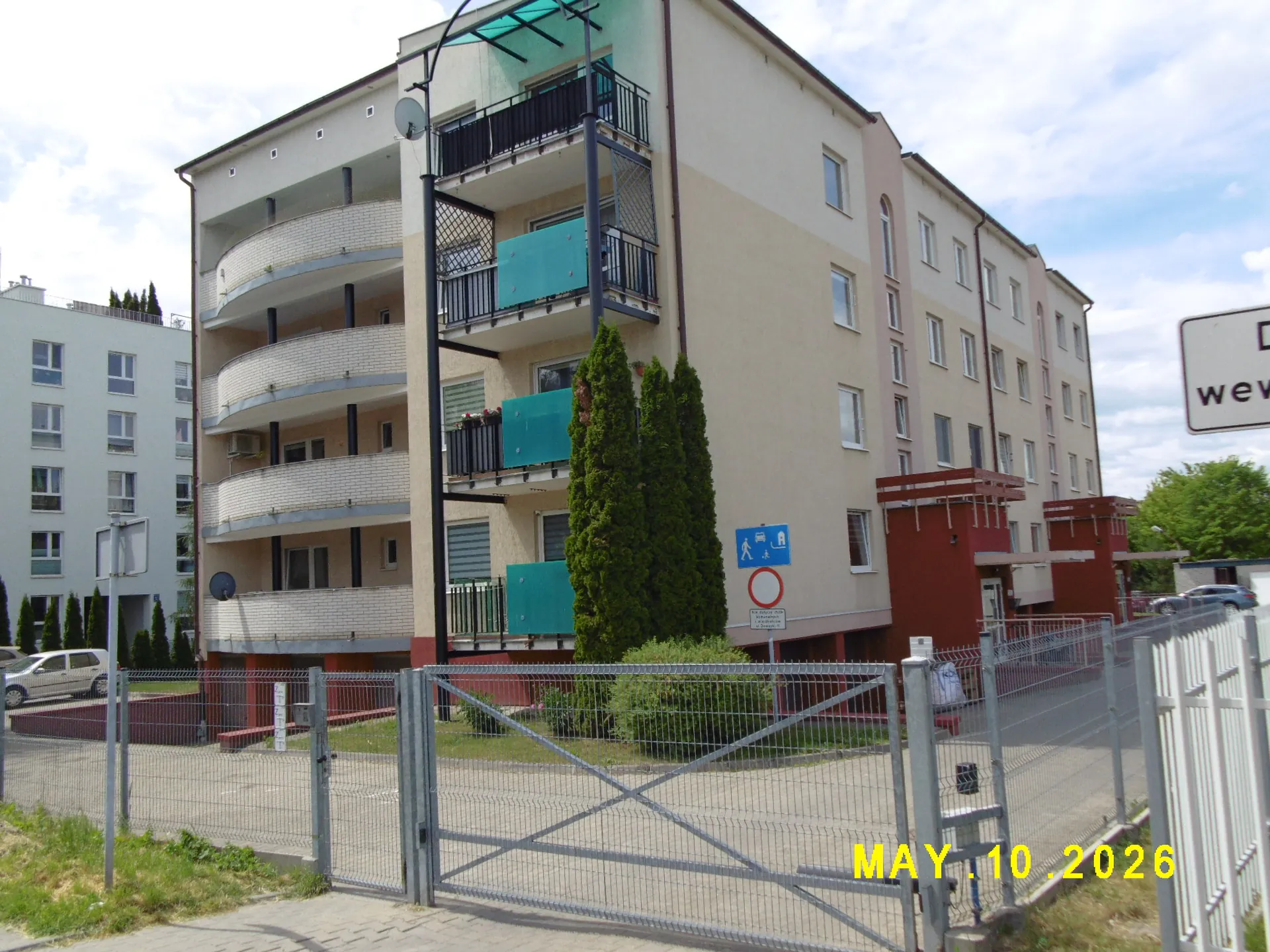 Wielopiętrowy budynek mieszkalny o jasnej elewacji z balkonami wyposażonymi w niebieskie i czarne barierki. Przed budynkiem znajduje się metalowy ogrodzenie z bramą oraz zielone krzewy i drzewka tujowe. Po prawej stronie widoczny jest fragment czerwonego przybudówki oraz znak drogowy. W prawym dolnym rogu zdjęcia znajduje się żółty napis z datą 'MAY 10 2026'.
