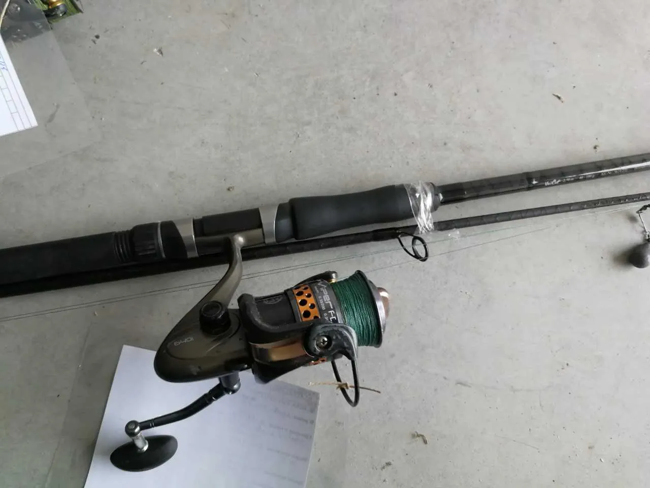 Widoczna wędkarska muszla z zielonym sznurem oraz czarny kij wędkarski leżące na szarym betonowym podłożu. Na muszli znajduje się napis 'VIPER'. W tle częściowo widoczny biały arkusz papieru z nieczytelnym tekstem oraz czarny przedmiot przypominający uchwyt. Obok kija wędkarskiego leży mały czarny pierścień.