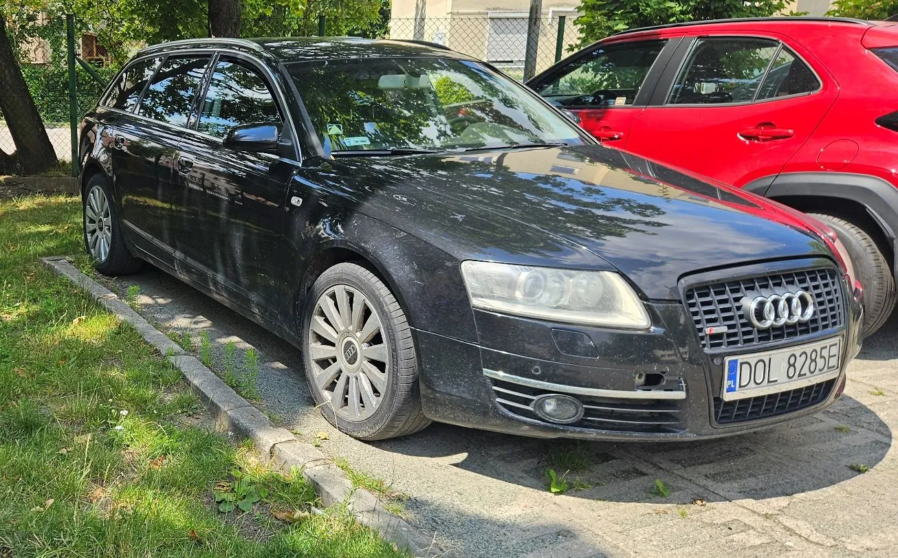 Czarny samochód marki Audi w nadwoziu kombi zaparkowany na poboczu obok krawężnika. Samochód ma polską tablicę rejestracyjną o numerze DOL 8285E. W tle widoczny jest fragment czerwonego samochodu oraz zielone drzewa i krzewy.