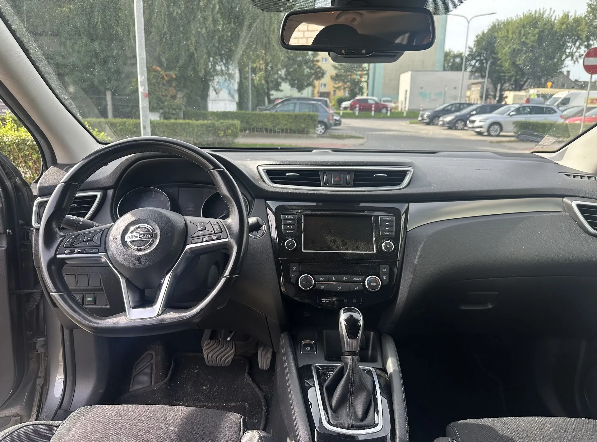 Widok z wnętrza samochodu marki Nissan, ukazujący deskę rozdzielczą, kierownicę z logo Nissan, konsolę centralną z ekranem i przyciskami oraz lewark zmiany biegów. Przez przednią szybę widoczny jest parking z zaparkowanymi samochodami, drzewa i budynki w oddali.