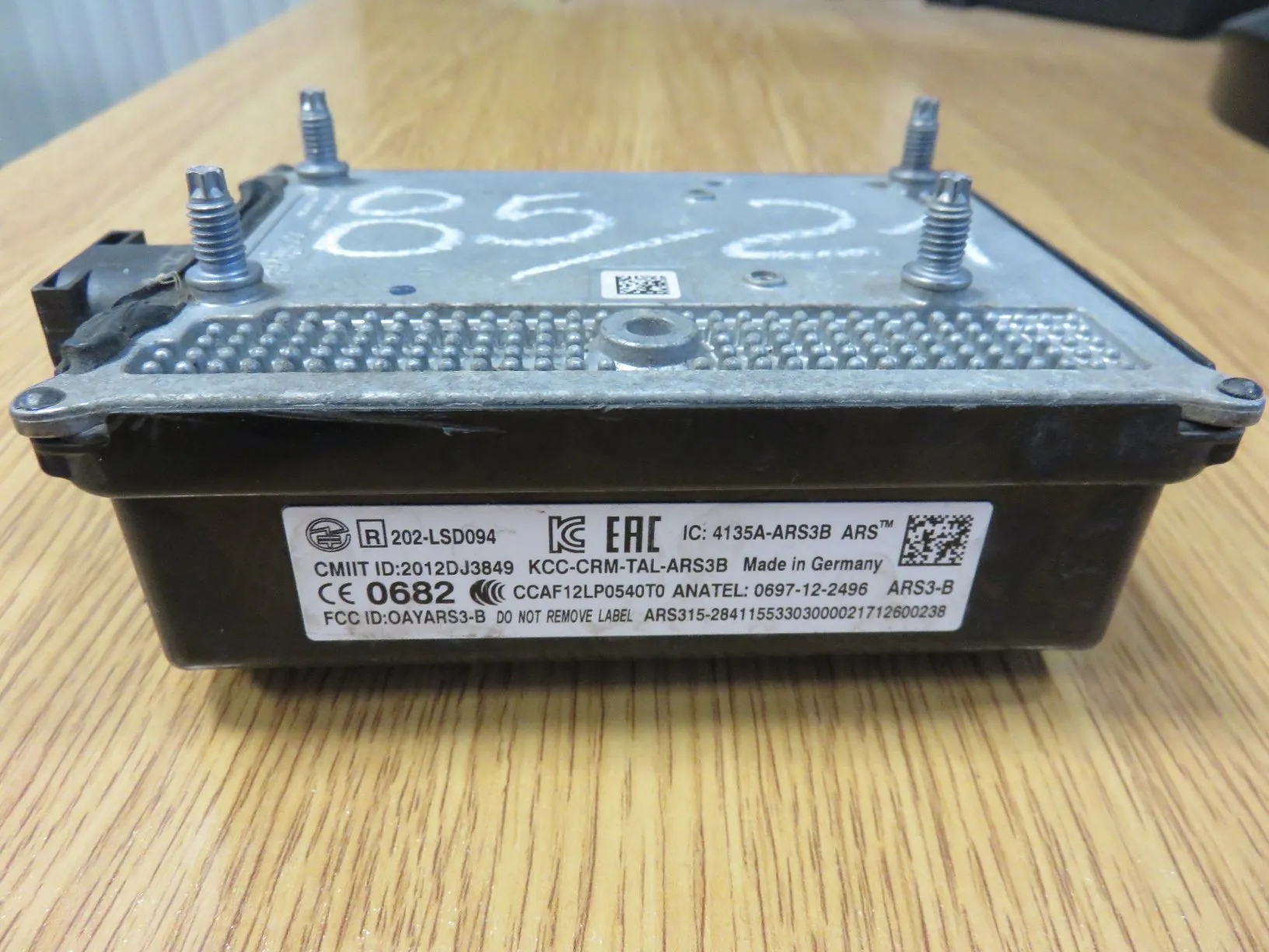 Srebrny, metalowy moduł elektroniczny z czarną obudową u dołu, leżący na drewnianym blacie. Na górnej części obudowy widnieją odręczne napisy markerem. Na przednim panelu znajduje się biała etykieta z licznymi oznaczeniami technicznymi i certyfikatami, w tym tekst: 'R 202-LSD094', 'EAC', 'IC: 4135A-ARS3B ARS™', 'CMIIT ID: 2012DJ3849', 'KCC-CRM-TAL-ARS3B Made in Germany', 'CE 0682', 'ANATEL: 0697-12-2496 ARS3-B', 'FCC ID: OAYARS3-B'. Urządzenie posiada cztery metalowe śruby mocujące w rogach.