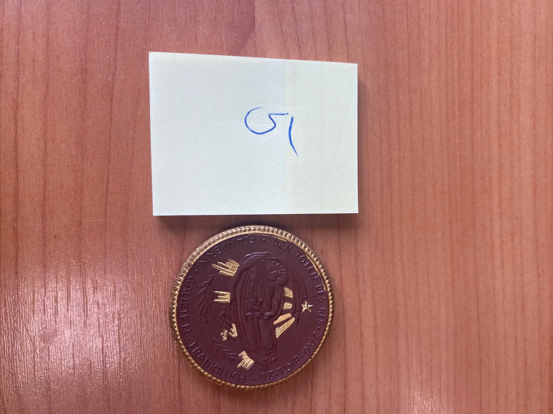 Na drewnianym tle leży biały kwadratowy kawałek papieru z niebieskim ręcznym napisem '51'. Poniżej znajduje się okrągły medal o brązowym kolorze z złotymi detalami. Medal posiada wygrawerowany napis: 'DEN TOTEN DES KRIEGES 1914-1918 FRANKREICH GALLIEN FLANDERN' oraz centralną postać z wieńcem i elementami dekoracyjnymi.