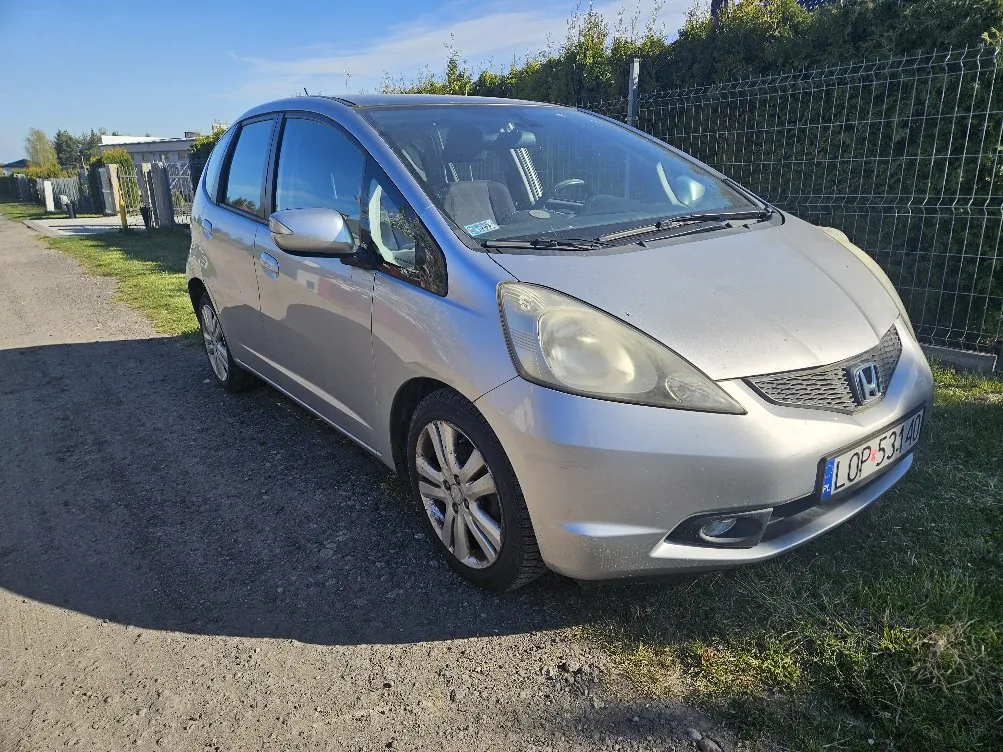 Srebrny samochód marki Honda zaparkowany na drodze żwirowej. Widoczna tablica rejestracyjna LOP 53140. W tle siatka ogrodzeniowa, zieleń i błękitne niebo. Samochód zwrócony lekko w prawo, widok z przodu i boku.