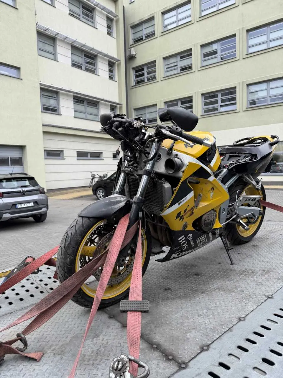 Żółto-czarny motocykl marki Kawasaki z widocznymi napisami 'BOS' i 'Kawasaki' na boku, zabezpieczony czerwonymi paskami na metalowej platformie z otworami. W tle budynek o jasnozielonej elewacji z oknami, zaparkowane samochody (w tym szary z tablicą rejestracyjną E5 4271) oraz rower. Motocykl ma żółte felgi i stoi na podstawce.