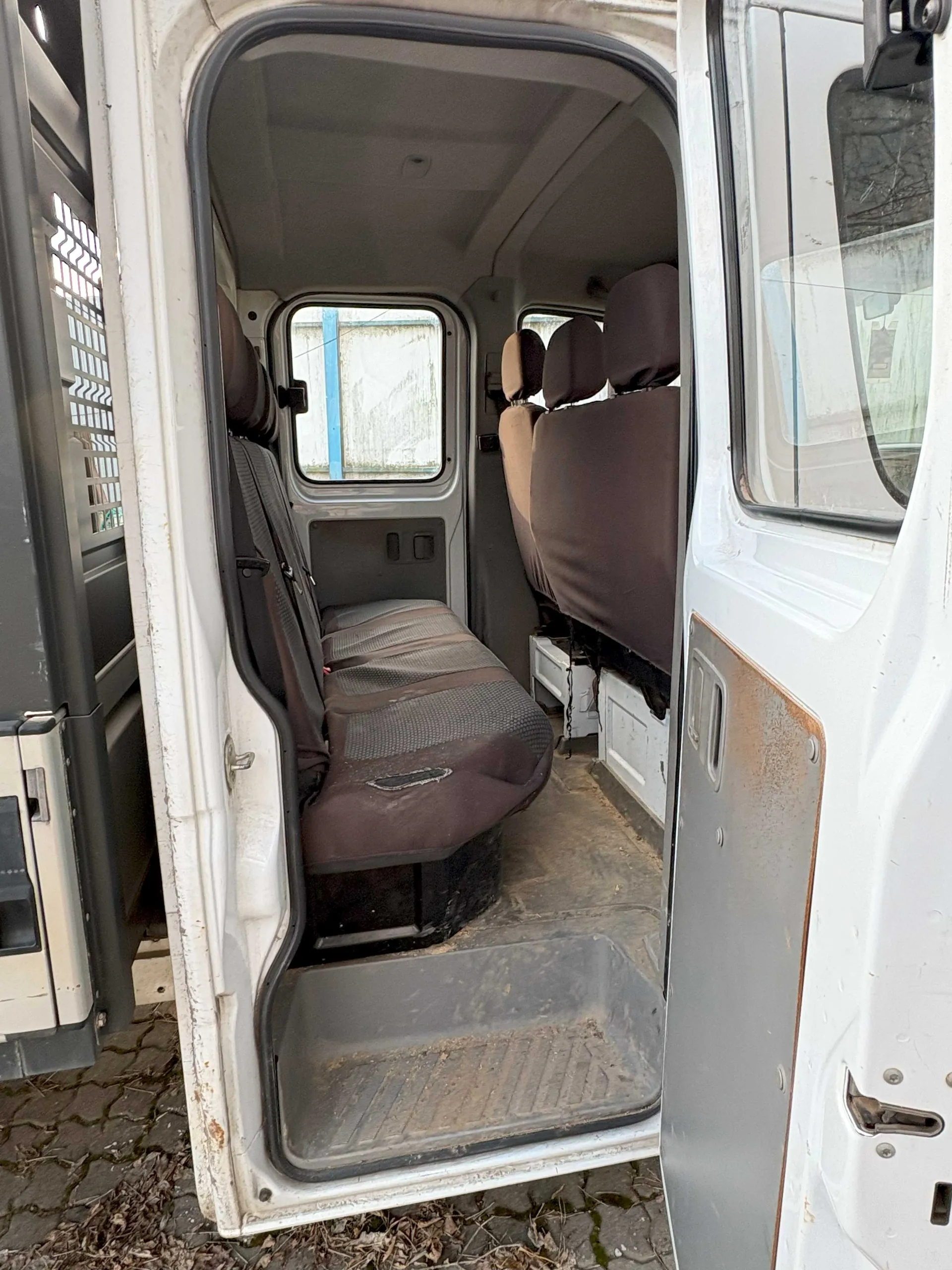 Wnętrze furgonetki z otwartymi drzwiami bocznymi. Widoczne są brązowe i szare siedzenia, zniszczona podłoga z brudem oraz zardzewiałe elementy drzwi. Po lewej stronie częściowo widoczny inny pojazd. Za oknem furgonetki budynek z niebieską kolumną i zasłoną. Podłoga zewnętrzna z bruku i suchymi liśćmi.