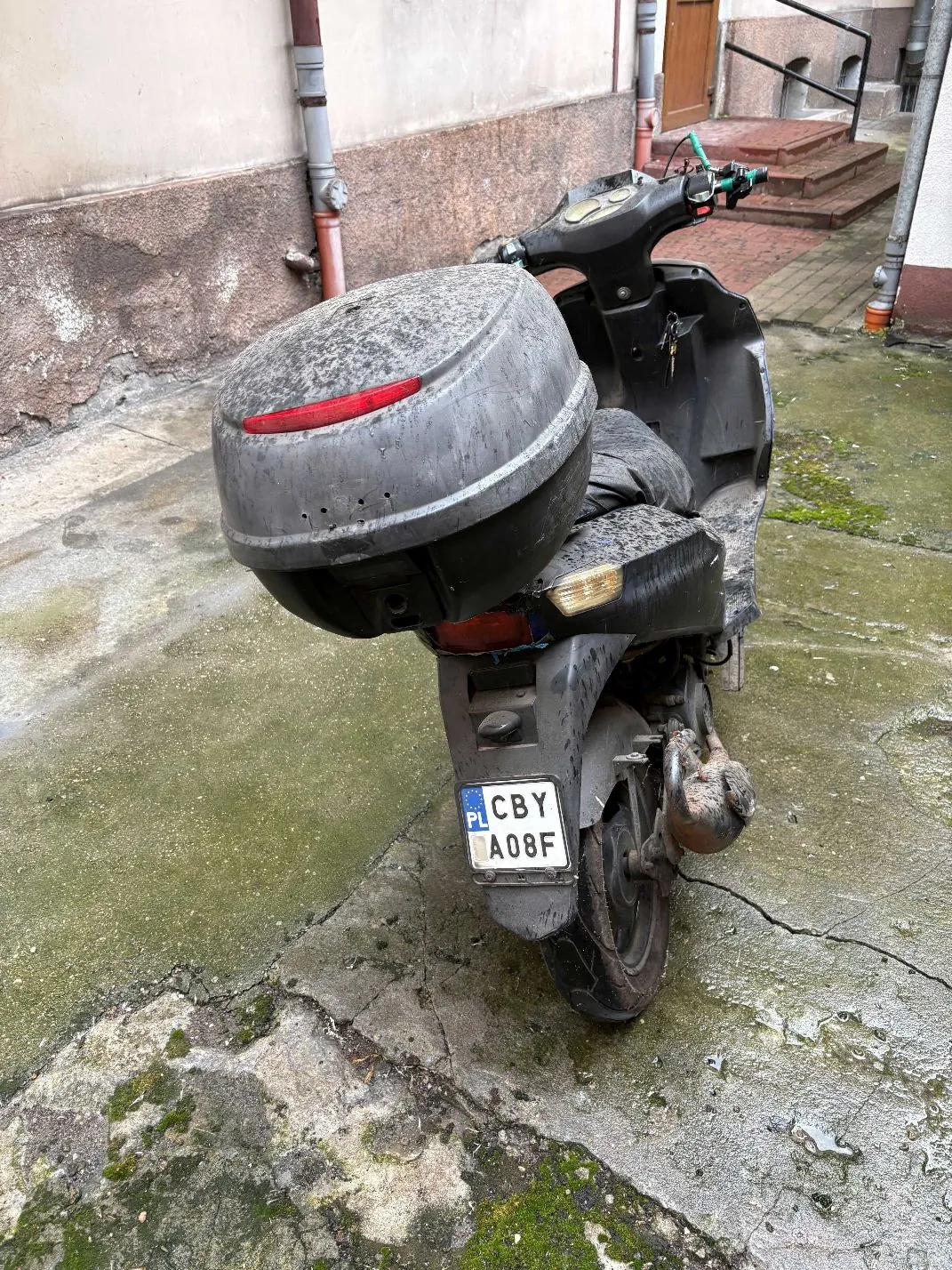 Zdjęcie z tyłu brudnego, czarnego skutera zaparkowanego na betonowym podłożu. Skuter posiada zamontowany duży, zakurzony kufer z czerwonym odblaskiem. Widoczna jest tablica rejestracyjna o treści 'CBY A08F' z oznaczeniem PL. W tle widać ścianę budynku oraz schody prowadzące do wejścia.