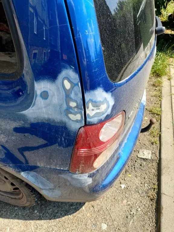 Zbliżenie na tylną, niebieską część nadwozia samochodu. Na karoserii, w okolicach tylnej lampy, widoczne są jasne plamy szpachli lub podkładu, wskazujące na przygotowanie powierzchni do naprawy lakierniczej. Widoczna jest czerwono-biała tylna lampa oraz fragment koła i tylnej szyby.