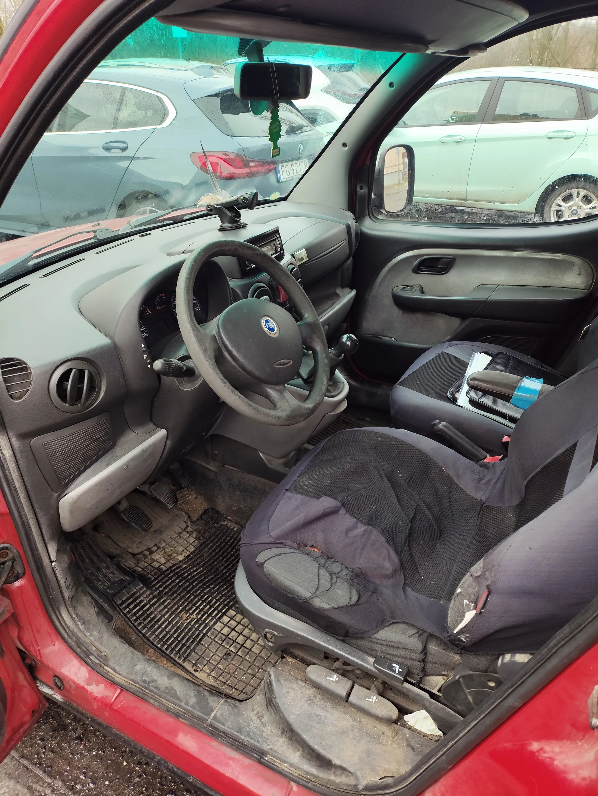 Wnętrze czerwonego samochodu z otwartą kierowcą drzwiami. Widoczna czarna kierownica z logo Fiata, szara deska rozdzielcza z licznikami, brudne dywaniki i pedały. Czarne siedzenia z widocznymi uszkodzeniami, na środkowej konsoli niebieska taśma. Przez przednią szybę widać zaparkowane samochody, w tym srebrny z tablicą rejestracyjną FG 9217P. W lusterku wstecznym wiszący zielony odświeżacz powietrza. Zewnętrzne tło z drzewami i innymi pojazdami.