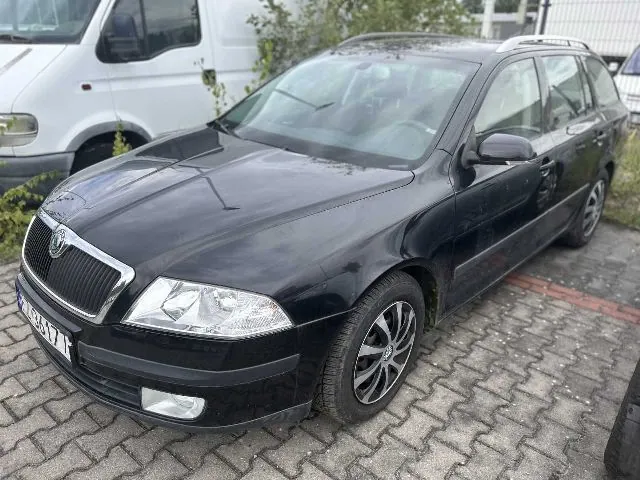 Czarny samochód osobowy marki Škoda w nadwoziu kombi, zaparkowany na kostce brukowej. Pojazd jest widoczny z przodu i z boku. W tle znajduje się biały samochód dostawczy.