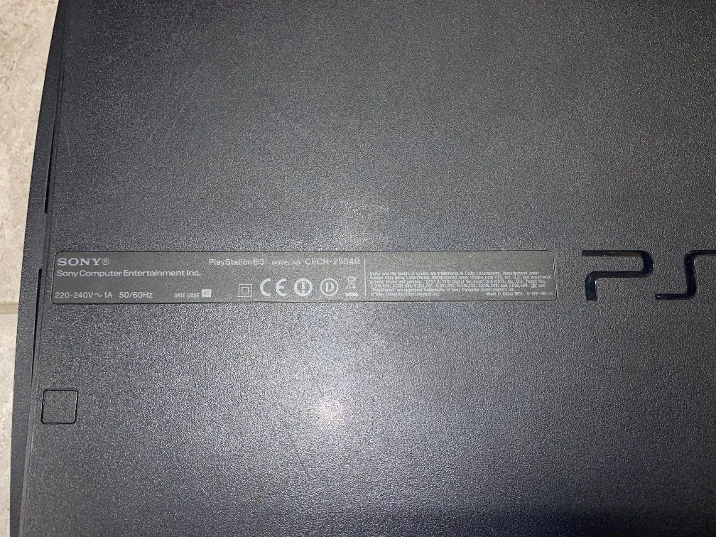 Zbliżenie na dolną część obudowy konsoli PlayStation 3 w kolorze czarnym. Widoczna jest srebrna tabliczka znamionowa z tekstem: 'SONY® Sony Computer Entertainment Inc. PlayStation®3 MODEL NO CECH-2504B 220-240V ~ 1A 50/60Hz'. Obok znajdują się symbole certyfikacji, w tym CE. Po prawej stronie widoczny jest fragment logo PS.