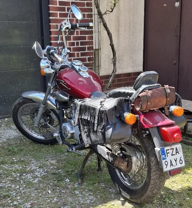 Czerwony motocykl zaparkowany na trawie i żwirze, z widoczną tablicą rejestracyjną "FZA 9AY6" (PL). Motocykl wyposażony w brązową skórzaną torbę na bagażniku i czarną torbę z frędzlami po boku. W tle ceglana ściana, drzewo i brązowe drzwi. Widoczne lusterka, pomarańczowe kierunkowskazy oraz chromowane elementy.