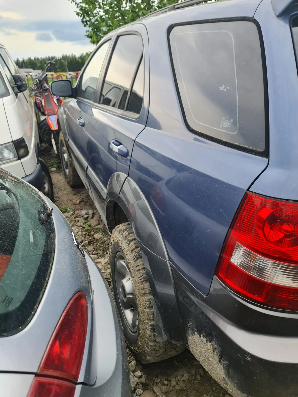 Zbliżenie na niebieski SUV zaparkowany na nierównym, kamienistym terenie. Po lewej stronie widoczne są części białego samochodu i srebrnego auta, a w tle czerwony skuter. Samochód ma brudne opony i zarysowane boki. W tle drzewa i chmurne niebo. Na tylnym oknie SUV widoczny jest niewielki, nieczytelny napis.
