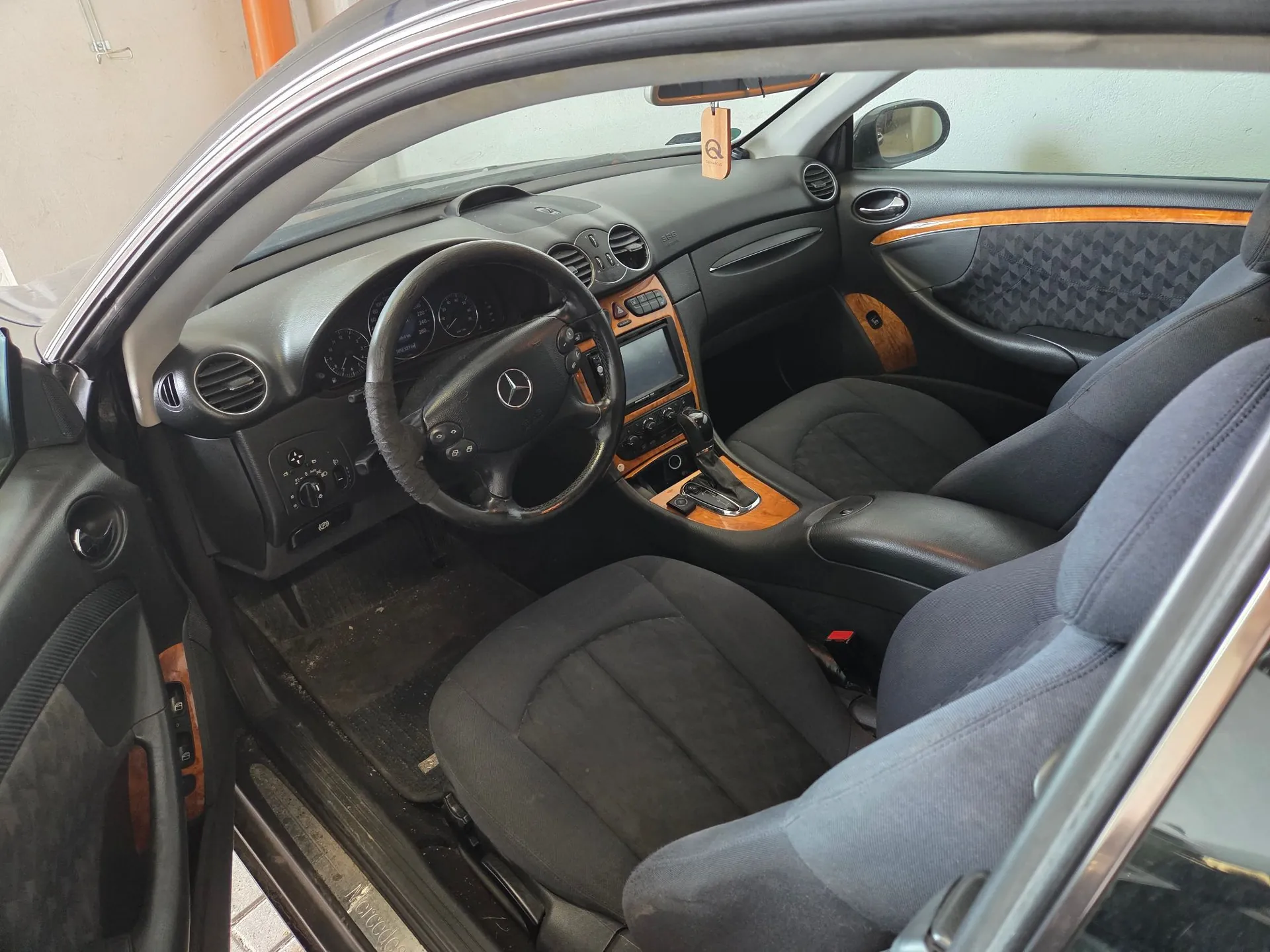 Wnętrze samochodu marki Mercedes-Benz widziane z perspektywy otwartych drzwi kierowcy. Widoczna jest czarna tapicerka materiałowa na fotelach, ciemna deska rozdzielcza z drewnianymi wstawkami oraz kierownica z logo marki w centrum. W konsoli środkowej znajduje się ekran multimedialny i lewarek zmiany biegów. Drzwi mają wykończenia z drewna i czarną tapicerkę z przeszyciami.