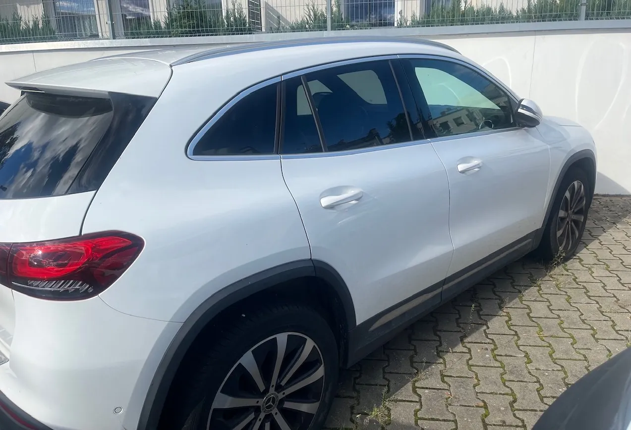 Boczny widok białego samochodu typu SUV zaparkowanego na kostce brukowej. Widoczna jest tylna część pojazdu z czerwonym reflektorem, srebrne klamki oraz ciemne szyby. W tle znajduje się biały mur i zielone krzewy za ogrodzeniem.