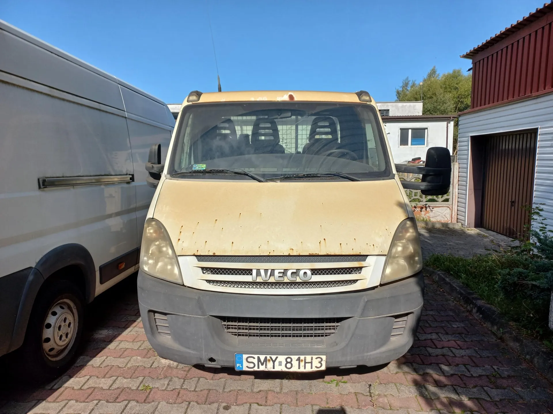 Zdjęcie przodu żółtego samochodu dostawczego marki Iveco zaparkowanego na kostce brukowej. Pojazd ma widoczne ślady zużycia i zabrudzenia na masce. Tablica rejestracyjna to SMY 81H3. Po lewej stronie widoczny jest fragment białego vana, a w tle jasny budynek z brązowym garażem i niebieskie niebo.