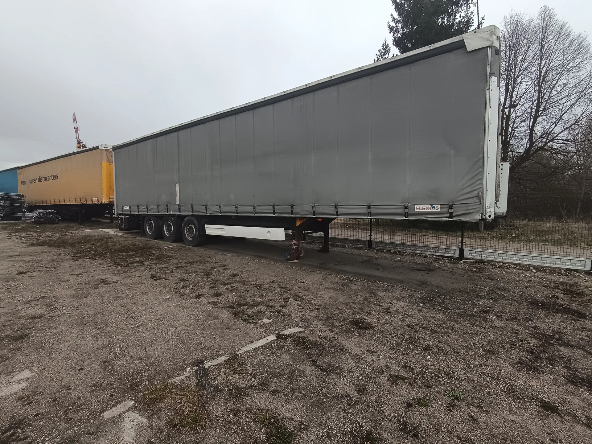 Dwa przyczepy ciężarowe zaparkowane na terenie z żwirem. Przyczepa na pierwszym planie jest szara z napisem 'FLEXOS' na boku, posiada trzy osie. W tle żółta przyczepa z częściowo widocznym napisem 'van wren distributers'. Wokół teren z rzadką trawą, metalowe ogrodzenie, drzewa bez liści i pochmurne niebo.