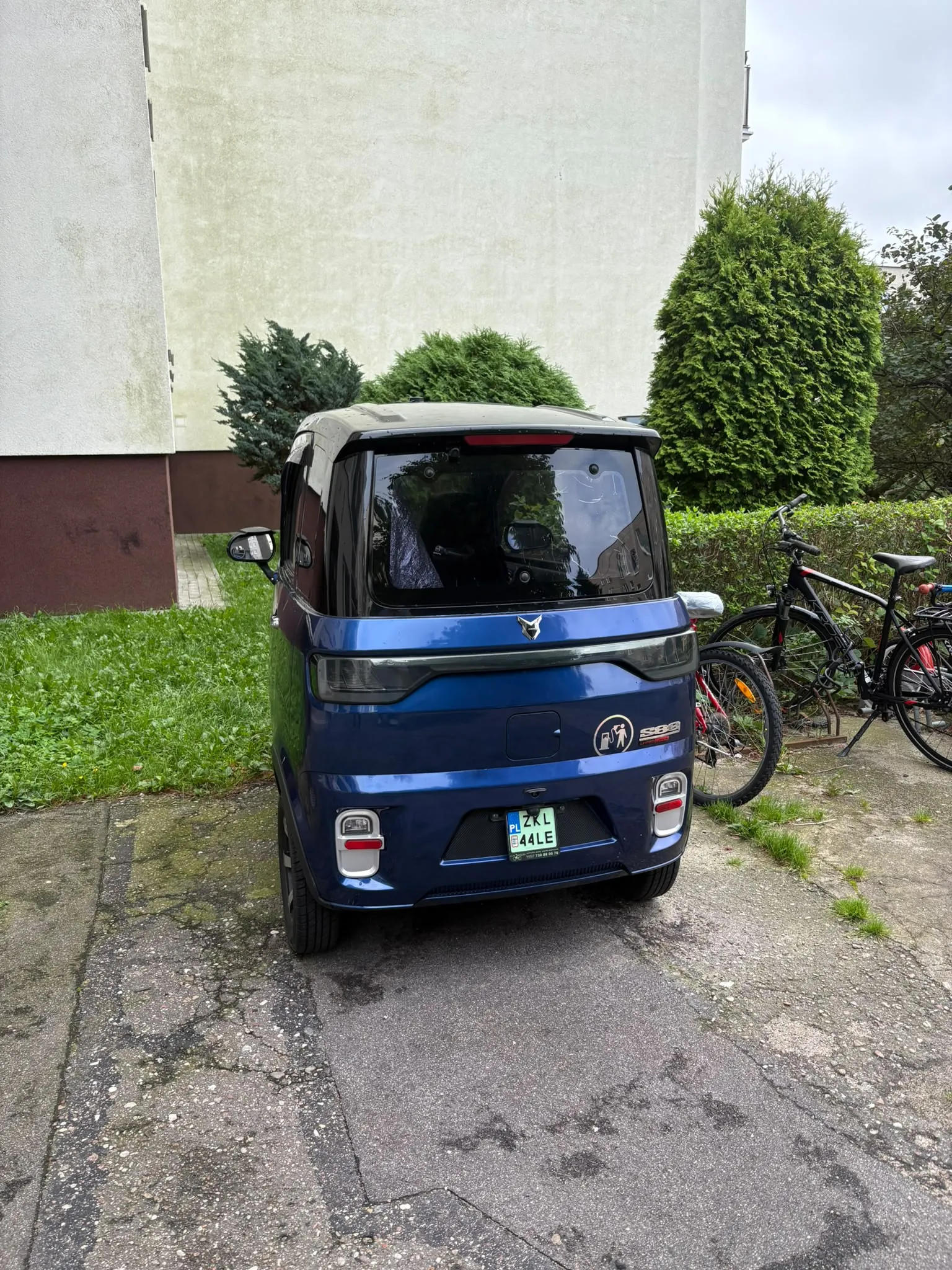 Niebieski, kompaktowy samochód elektryczny zaparkowany tyłem na betonowym podjeździe. Pojazd ma charakterystyczny, kanciasty kształt i rejestrację o treści ZKL 44LE. W tle widoczna jest jasna ściana budynku, zielone krzewy oraz dwa rowery zaparkowane po prawej stronie.
