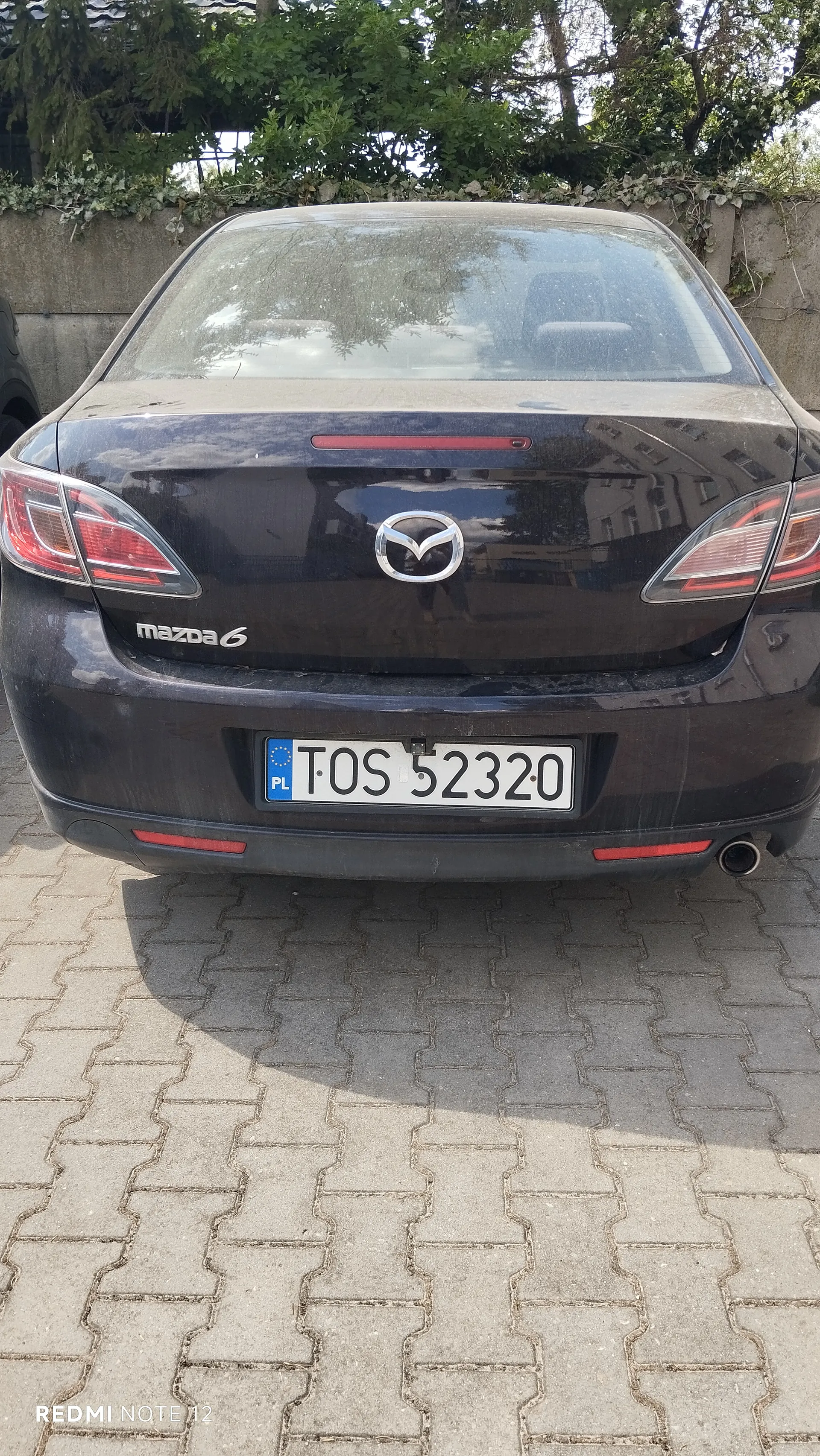 Tył czarnego samochodu Mazda 6 zaparkowanego na brukowanej powierzchni. Widoczny jest biały tabliczka rejestracyjna z niebieskim pasem z napisem PL oraz czarnymi literami i cyframi TOS 52320. Na bagażniku umieszczono srebrny emblemat marki i napis mazda 6. W tle betonowy mur z zielonymi pnączami oraz drzewa. Na dole zdjęcia widoczny napis REDMI NOTE 12.