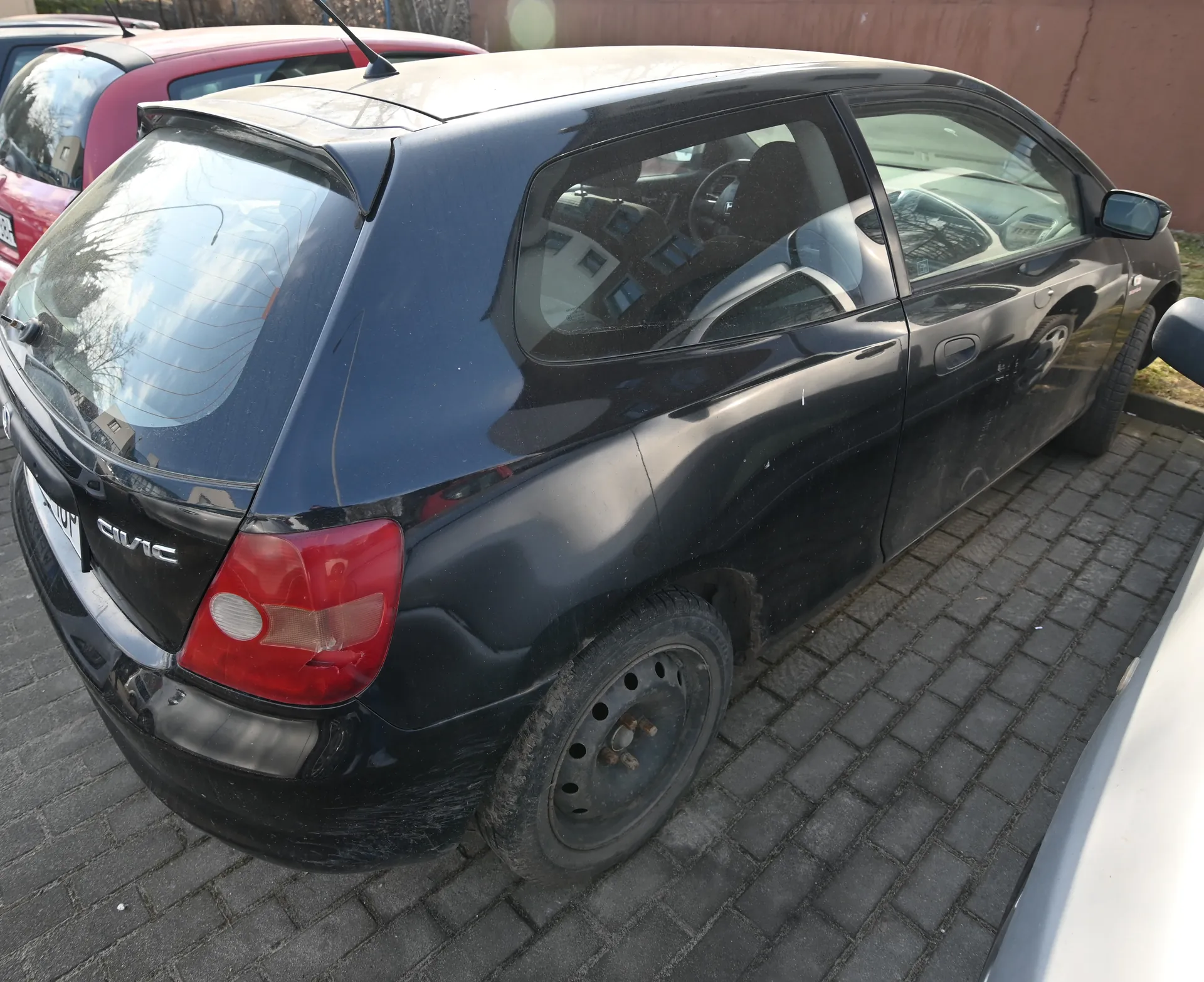 Czarny samochód Honda Civic widziany od tyłu, zaparkowany na brukowanej powierzchni. Na bagażniku widoczny napis CIVIC, czerwone światło tylnego reflektora oraz tablica rejestracyjna. W tle czerwony i biały samochód oraz brązowa ściana. Samochód ma brakującą osłonę felgi na tylnej oponie. Szkła samochodu odbijają otoczenie.