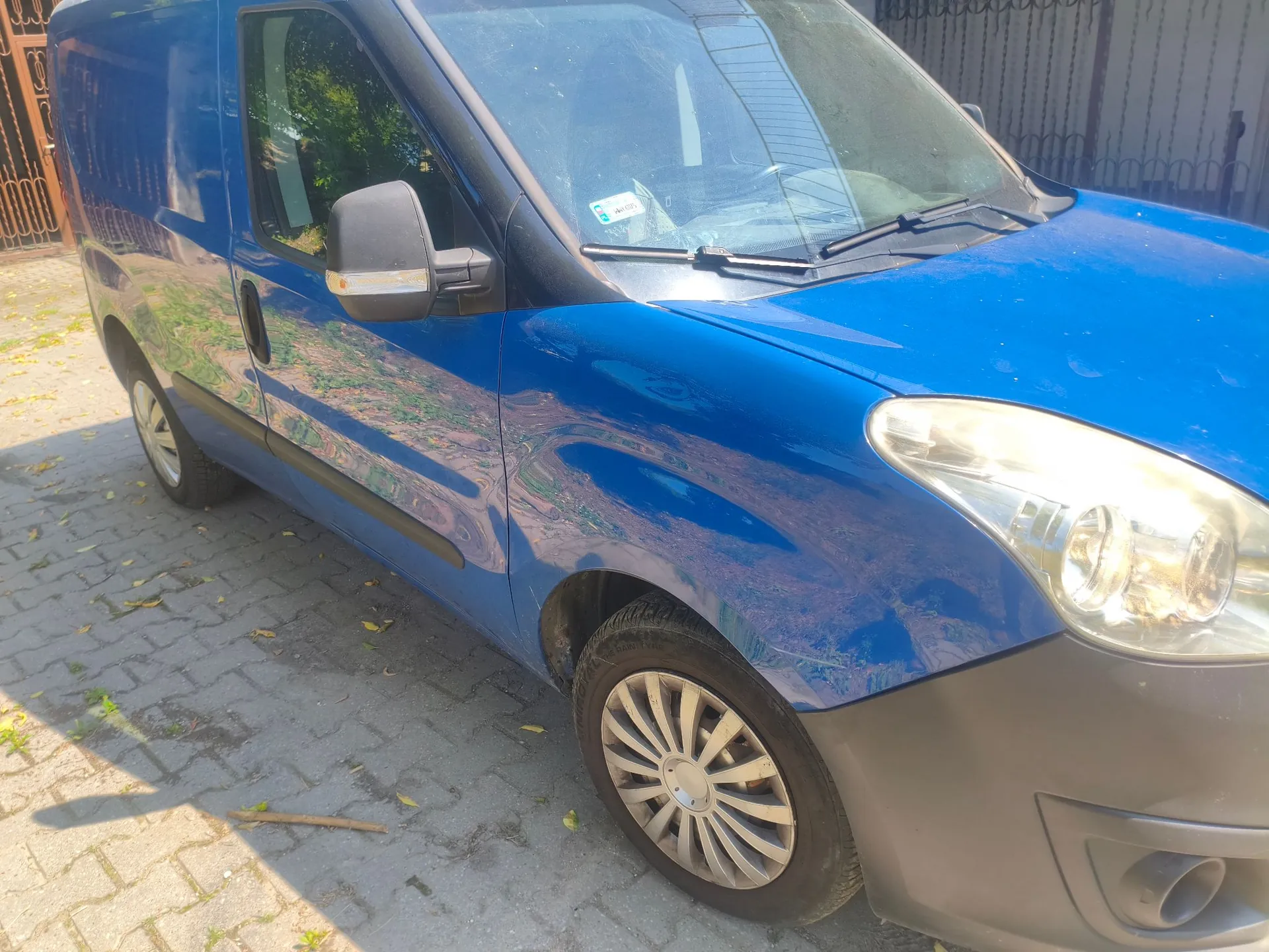 Niebieski samochód dostawczy zaparkowany na kostce brukowej. Widoczny jest przód i bok pojazdu, w tym prawe przednie koło z kołpakiem, reflektor oraz lusterko boczne. W tle widać metalowy płot i zieleń drzew.