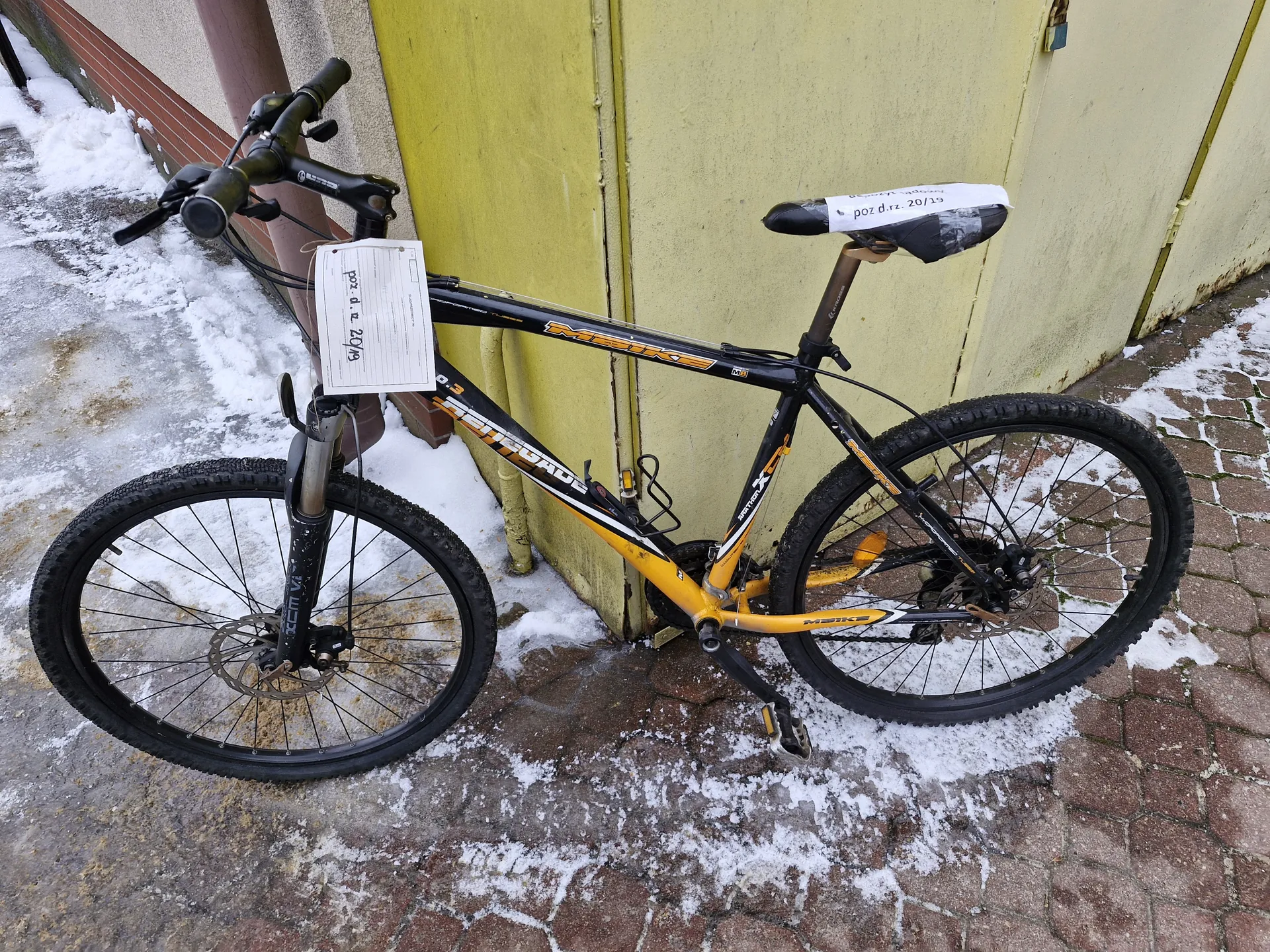 Czarno-żółty rower górski oparty o żółtą ścianę. Na kierownicy i siedzisku widoczne białe kartki z napisem "poz d.rz. 20/19". Podłoże z ceglanego chodnika częściowo pokryte śniegiem i lodem. W tle fragment budynku z brązowym rurem i śniegiem przy ścianie.