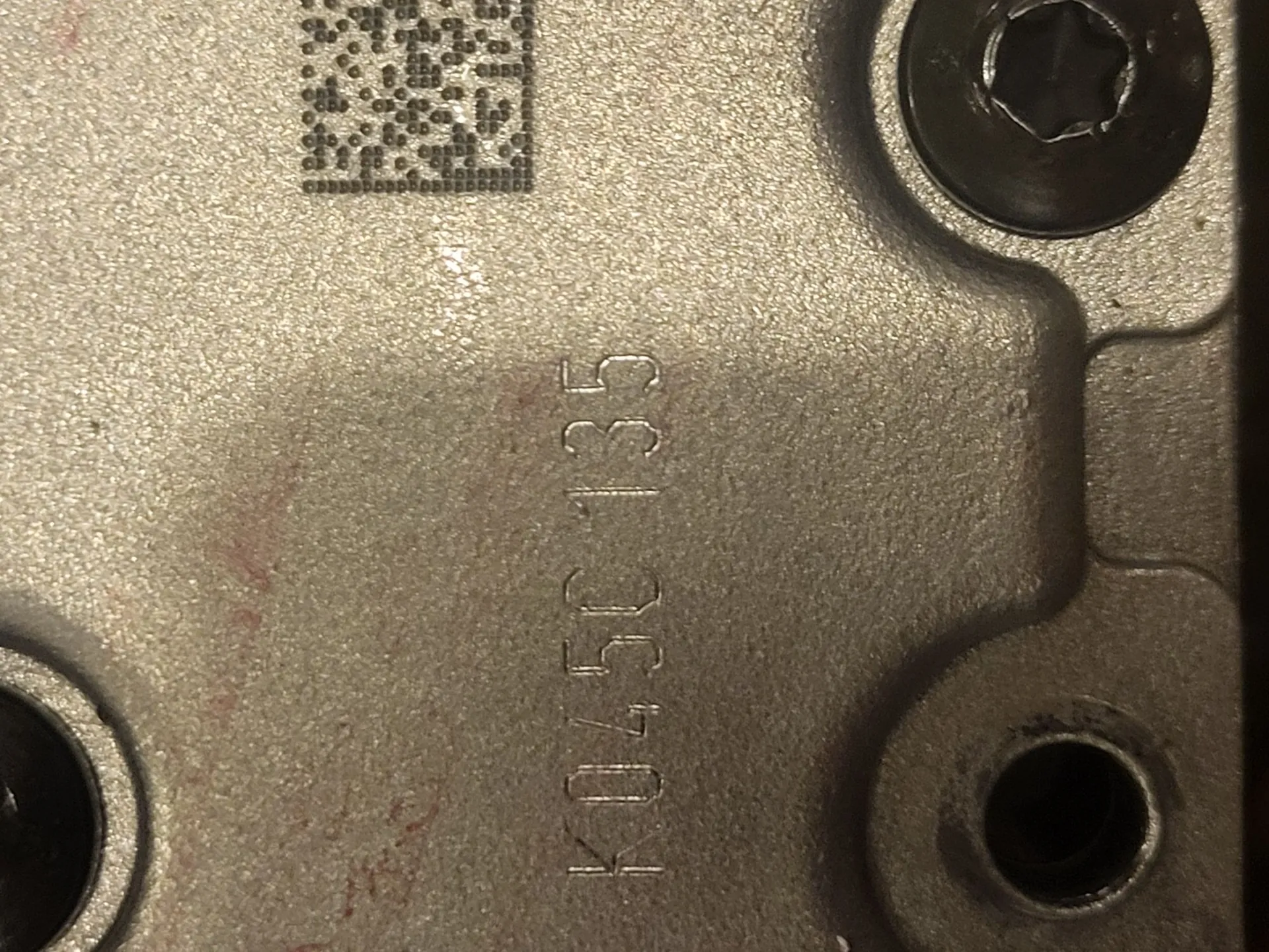 Zbliżenie na metalową, srebrną powierzchnię części mechanicznej. Na obrazie widoczny jest kod QR w górnej części oraz pionowy napis wyryty w metalu: 'K0450 C 135'. W prawym górnym rogu znajduje się czarna śruba z gniazdem imbusowym, a w dolnych rogach widoczne są otwory montażowe.