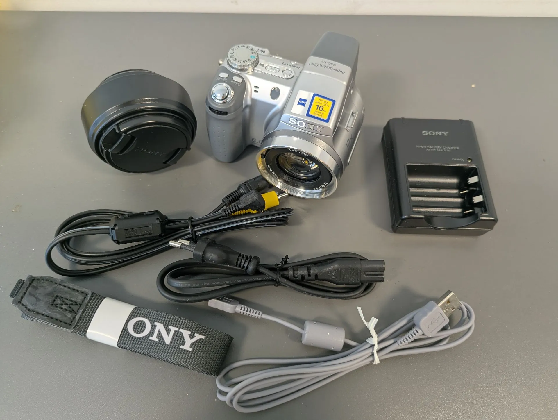 Zestaw akcesoriów fotograficznych ułożonych na szarym tle. W centrum znajduje się srebrny aparat cyfrowy Sony z obiektywem, na którym widnieje żółta naklejka z liczbą 16. Obok aparatu leży czarna osłona obiektywu, czarna ładowarka do baterii z napisem SONY oraz dwa czarne kable zasilające. Na dole znajduje się szary pasek z napisem ONY oraz szary kabel USB.