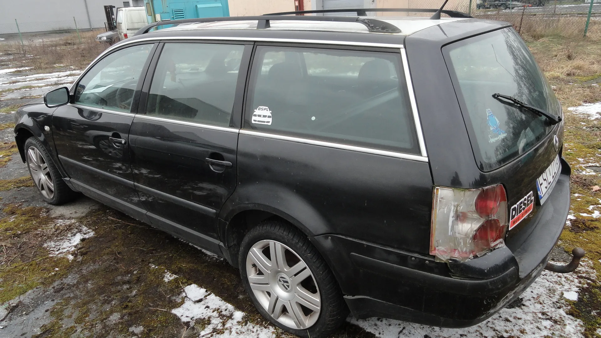 Czarny samochód typu kombi zaparkowany na terenie z zielonym mchem i resztkami śniegu. Samochód posiada dachowy bagażnik, srebrne felgi oraz naklejki na tylnym szybie, w tym czerwoną z napisem DIESEL. Widoczna jest tablica rejestracyjna z częściowo zasłoniętym numerem. W tle budynek z niebieską bramą i inny pojazd. Na bocznej szybie naklejka z napisem 85PL oraz ikoną autobusu.