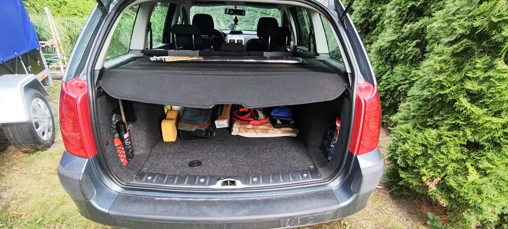 Otwarty bagażnik ciemnoszarego samochodu typu kombi. Wewnątrz znajdują się różne przedmioty: żółty kanister, czarna torba, czerwony kabel zwinięty w kable oraz inne drobne akcesoria. W tle widoczne są zielone krzewy oraz fragment niebieskiej plandeki po lewej stronie.