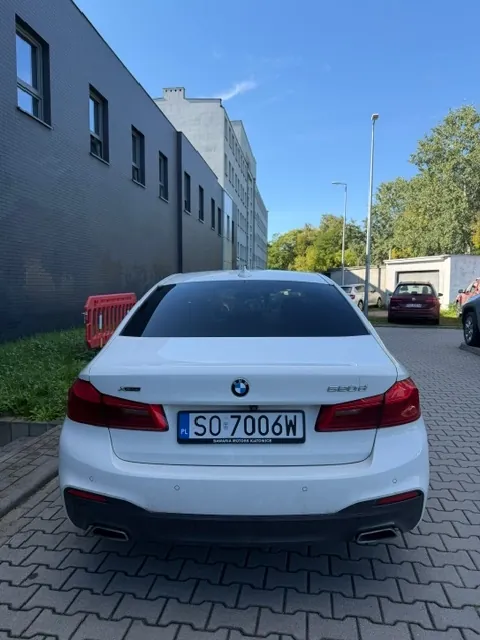 Widok z tyłu białego samochodu marki BMW model 520d zaparkowanego na kostce brukowej. Samochód ma tablicę rejestracyjną o numerze SO 7006W. W tle znajduje się ciemnoszary budynek z rzędem okien, zielona trawa oraz inne zaparkowane samochody pod jasnym, niebieskim niebem.