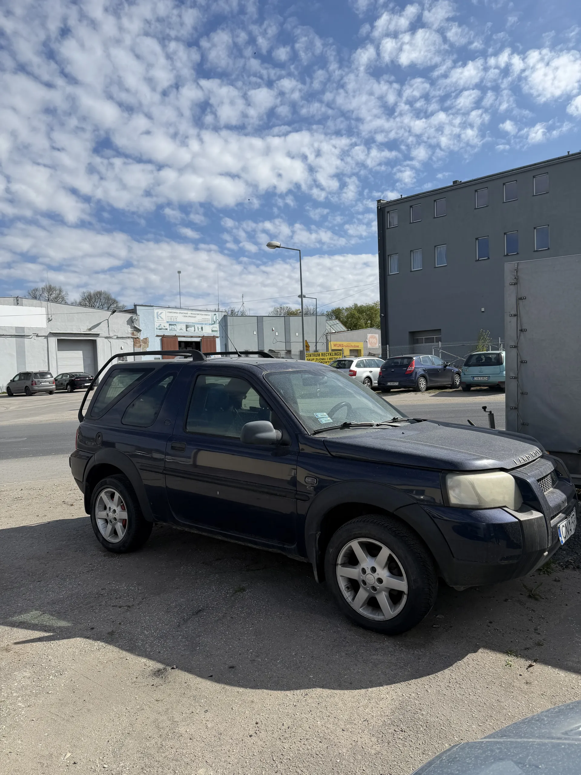 Ciemnoniebieski samochód zaparkowany na asfaltowej powierzchni po prawej stronie obrazu. Po lewej stronie widoczny jest szary budynek z oknami, a także niebo z białymi chmurami. W tle znajdują się inne zaparkowane samochody, latarnia uliczna oraz budynki przemysłowe.
