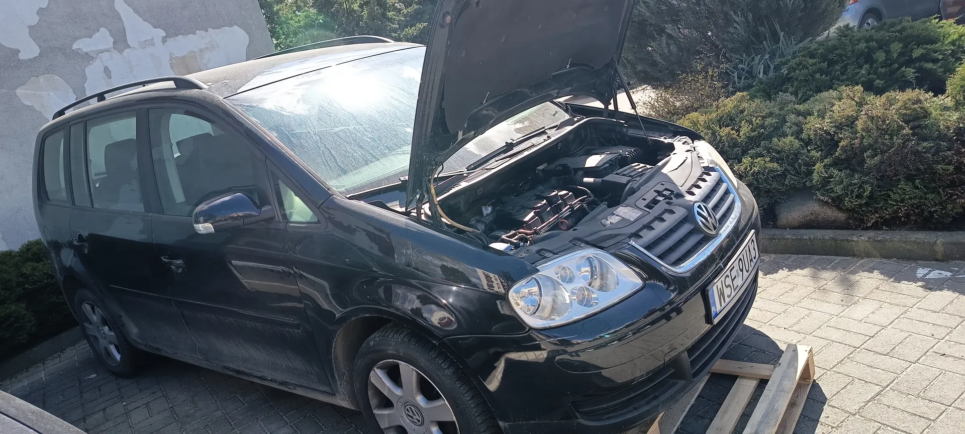 Czarny samochód Volkswagen z otwartą maską silnika, zaparkowany na brukowanej powierzchni. Widoczna tablica rejestracyjna WSE 9U43, przednie koło prawe oparte na drewnianym palecie. W tle zielone krzewy, fragment ściany z obłupioną farbą oraz drzewa. Samochód posiada dachowy bagażnik i lusterka boczne. Słoneczne oświetlenie odbija się w szybie i karoserii.