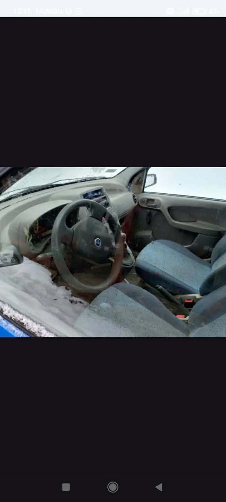 Wnętrze samochodu z widocznym kierownicą, uszkodzonym deski rozdzielczej i śniegiem na siedzeniach oraz podłodze. Siedzenia są niebieskie z wzorem, drzwi pasażerskie są otwarte, a na zewnątrz widoczny jest śnieg. W tle przez przednią szybę widać zimowy krajobraz.