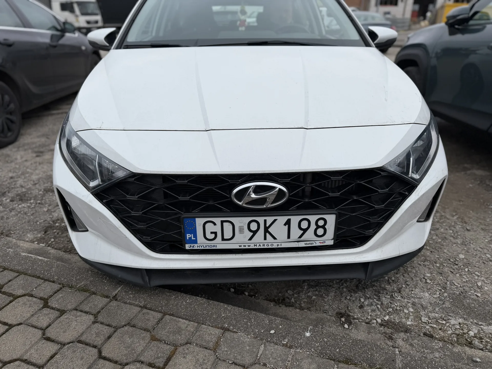 Biały samochód marki Hyundai z przodu, z widoczną tablicą rejestracyjną PL GD 9K 198. Na tablicy widoczne są dodatkowe napisy: HYUNDAI, www.MARGO.pl oraz logo Margo. Samochód stoi na brukowanej powierzchni, w tle znajdują się inne pojazdy i budynki.