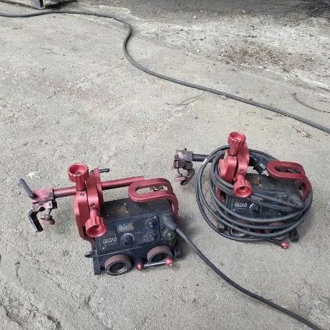 Dwa czerwone urządzenia techniczne, prawdopodobnie uchwyty lub stabilizatory z zamontowanymi czarnymi modułami elektronicznymi, leżą na betonowym podłożu. Urządzenia są połączone czarnymi kablami, z których jeden ciągnie się w stronę górnej krawędzi zdjęcia. Na obudowach modułów widnieje napis 'GE160'.