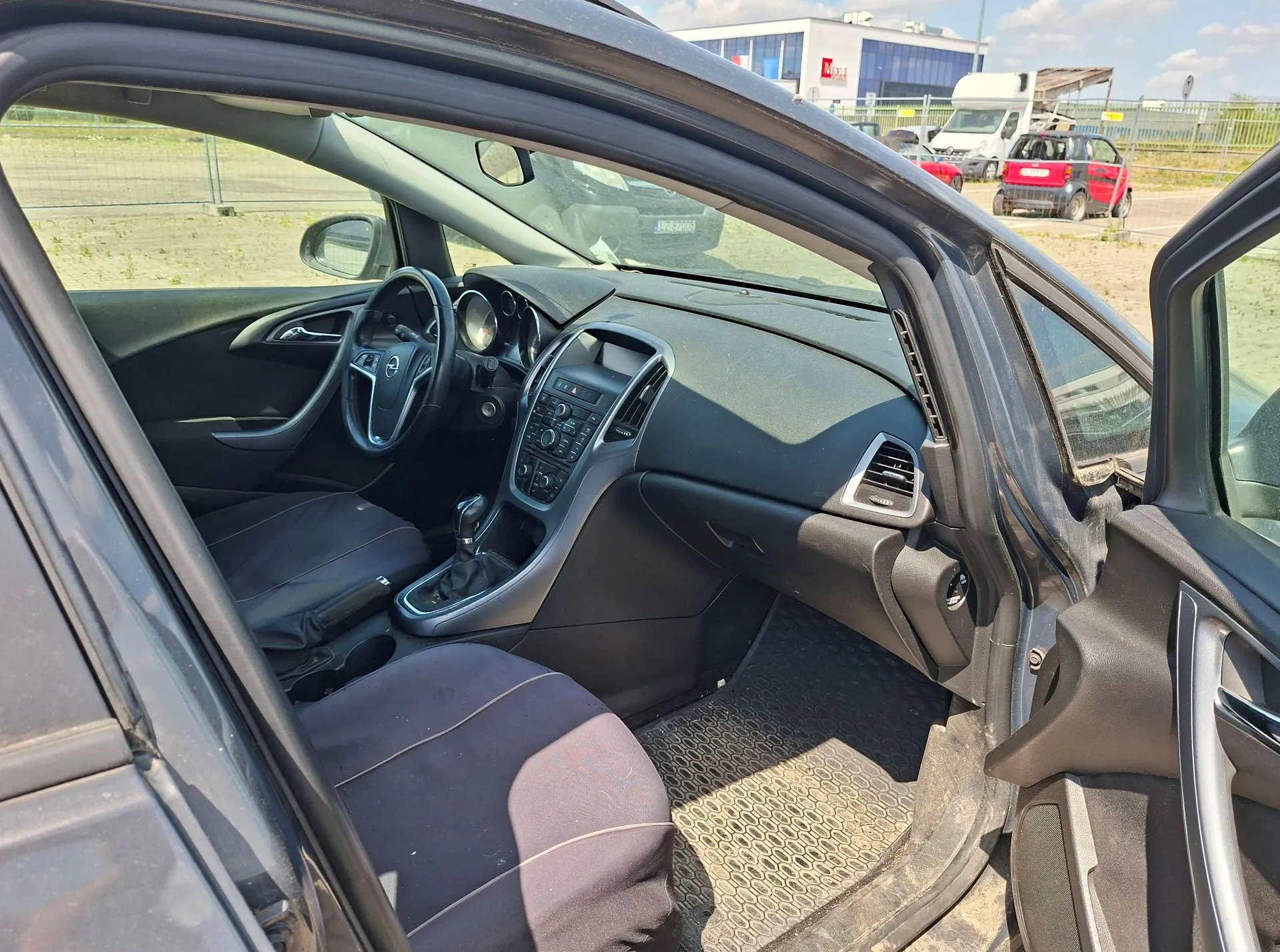 Wnętrze samochodu widziane z otwartych drzwi pasażera. Widoczna jest czarna deska rozdzielcza, kierownica z logo marki Opel, konsola środkowa z przyciskami i ekranem oraz ciemne fotele. Na podłodze znajduje się gumowa mata. Przez szyby widoczny jest parking z innymi samochodami i budynkiem w tle.