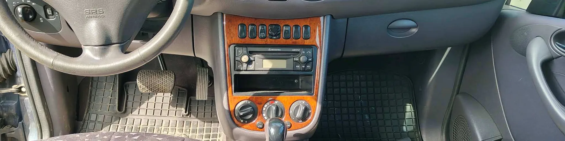 Wnętrze samochodu z widocznym kierownicą z napisem SRS AIRBAG, deską rozdzielczą z drewnianą obudową, odtwarzaczem CD, przyciskami sterującymi, gałką zmiany biegów, pedałami oraz czarnymi gumowymi dywanikami na podłodze. Kierownica znajduje się po lewej stronie, a deska rozdzielcza posiada przyciski i pokrętła klimatyzacji.