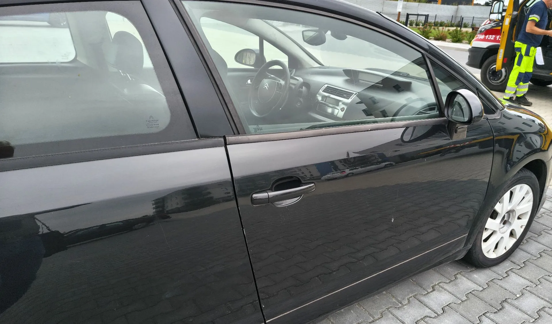 Zbliżenie na prawy bok czarnego samochodu osobowego zaparkowanego na kostce brukowej. Widoczne są przednie i tylne drzwi, lusterko boczne oraz przednie koło z jasną felgą. Przez szyby widać wnętrze pojazdu, w tym kierownicę i fotele. W tle, po prawej stronie, widoczny jest fragment żółtego pojazdu technicznego oraz osoba w odblaskowej kamizelce.