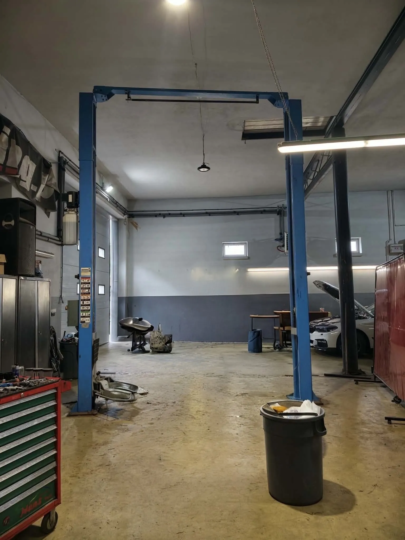Wnętrze warsztatu samochodowego z niebieską, dwukolumnową windą hydrauliczną w centrum. Po lewej stronie znajduje się czerwony wózek narzędziowy z szufladami oraz szafki metalowe. Po prawej stronie widoczny jest biały samochód z otwartą maską, stół warsztatowy oraz czerwona szafa. Na podłodze stoi czarny kosz na śmieci oraz leżą rozproszone części samochodowe. Pomieszczenie jest oświetlone lampami sufitowymi, a ściany są pomalowane na biało z szarym pasem w dolnej części.
