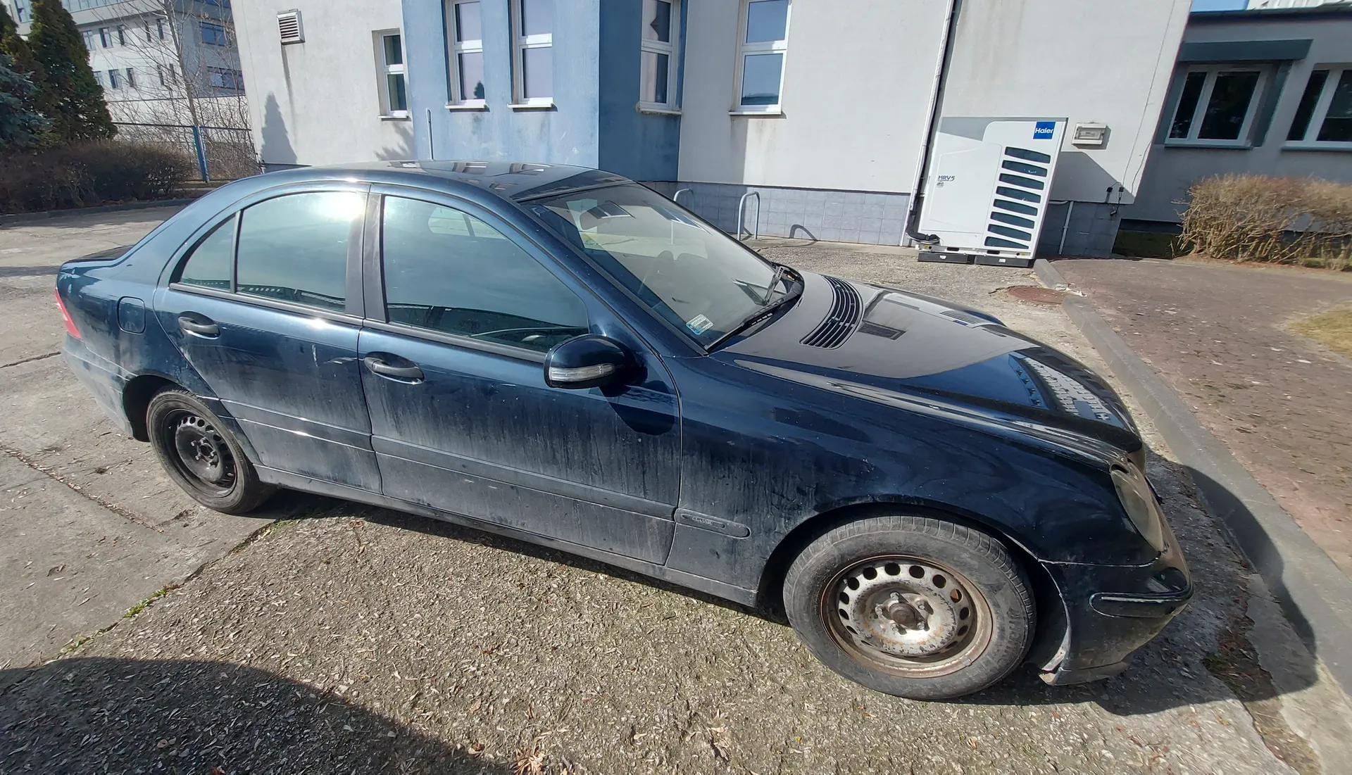 Ciemnoniebieski samochód osobowy typu sedan zaparkowany na podwórku z żwirem. Pojazd jest zabrudzony, z widocznymi śladami kurzu na karoserii i zardzewiałymi felgami. W tle budynek z białymi i niebieskimi ścianami, oknami oraz białą jednostką klimatyzacyjną z napisem "Haier". Obok budynku rosną krzewy i widoczne są inne zabudowania.