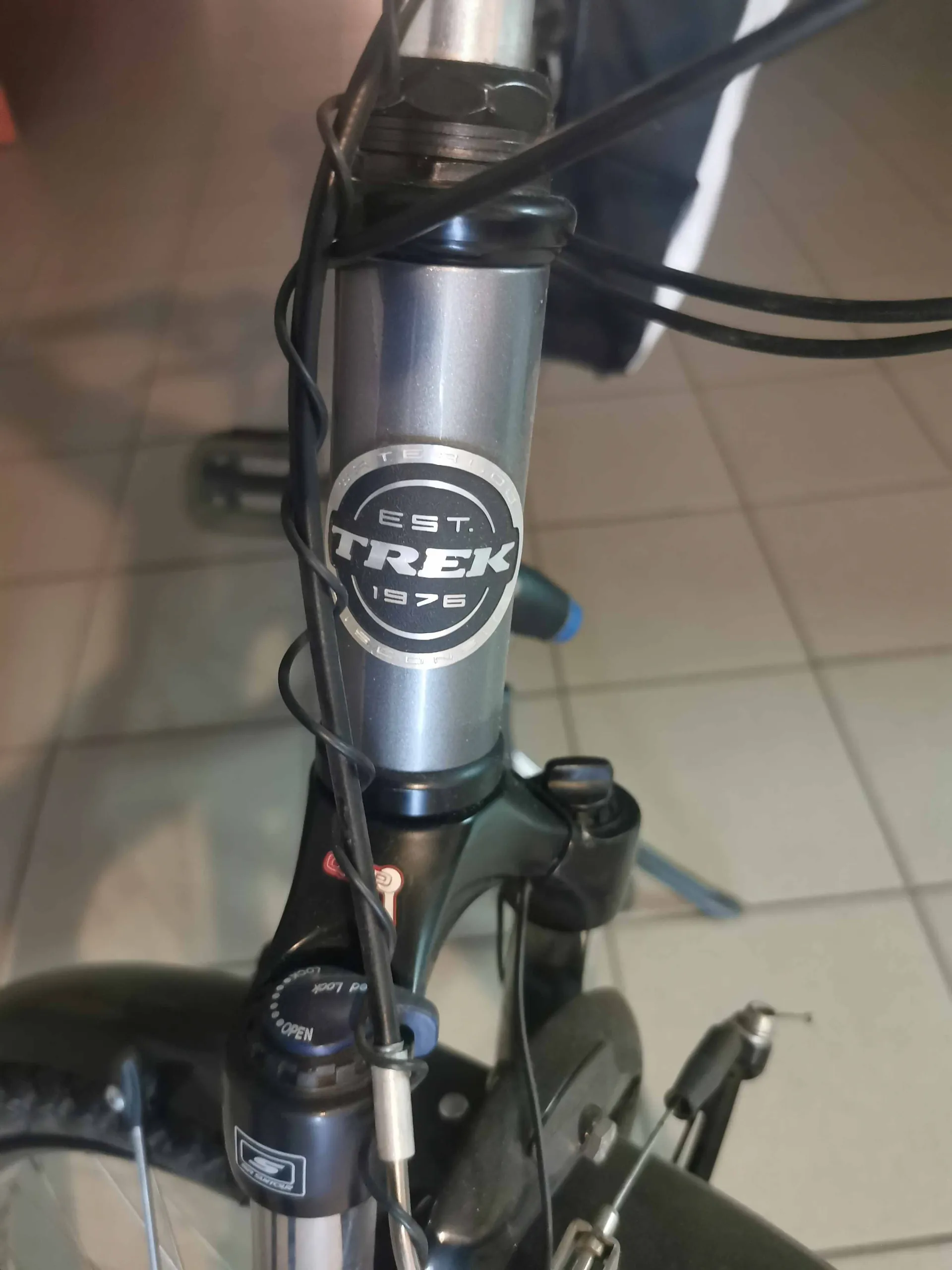 Zbliżenie na srebrną rurę kierownicy roweru marki Trek. Na rurze znajduje się okrągłe logo z napisem „EST. TREK 1976”. Wokół rury przebiegają czarne kable hamulcowe i przerzutek. W dolnej części widoczny jest czarny mostek kierownicy oraz fragment amortyzatora z napisem „Lock / Open”.