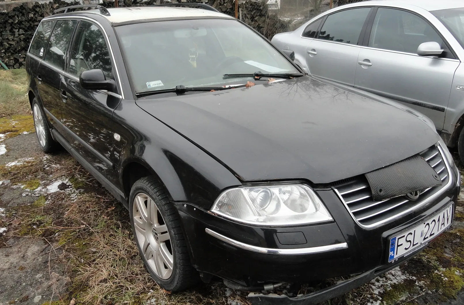 Czarny samochód typu kombi zaparkowany na terenie z suchą trawą i resztkami śniegu. Po prawej stronie widoczny srebrny samochód osobowy. Na czarnym samochodzie tablica rejestracyjna z napisem FSL 221AV oraz oznaczeniem PL. W tle ogrodzenie z drewnianymi bali i drzewa. Samochód posiada dachówkę i srebrne felgi.