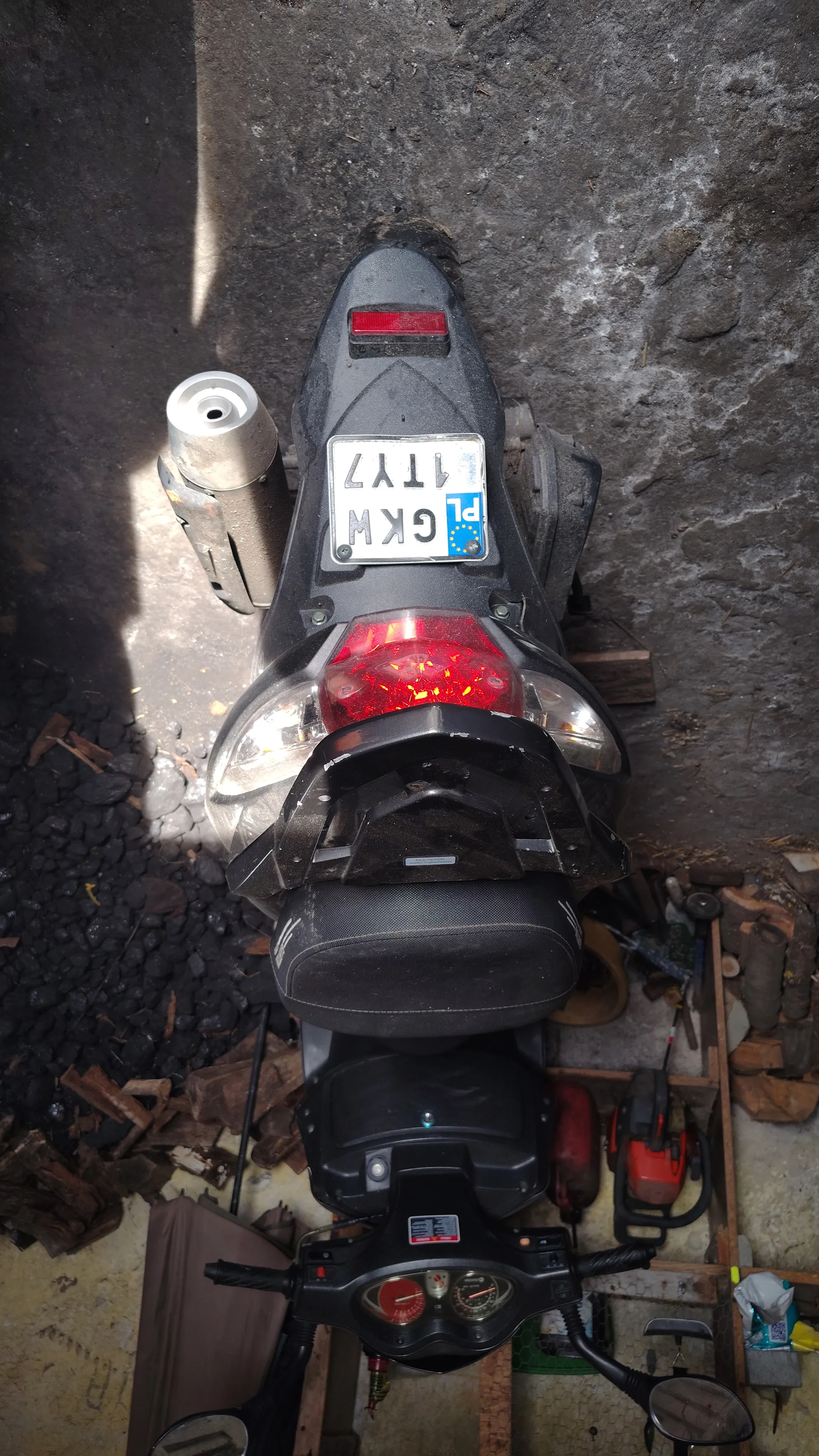 Czarny skuter widziany z góry, z tablicą rejestracyjną PL GKW 1TY7 umieszczoną pionowo. Skuter posiada czerwone światło tylne, wydech, kierownicę z lusterkami i wskaźnikami. W tle widoczne są węgiel, deski drewniane, narzędzia oraz betonowa podłoga.
