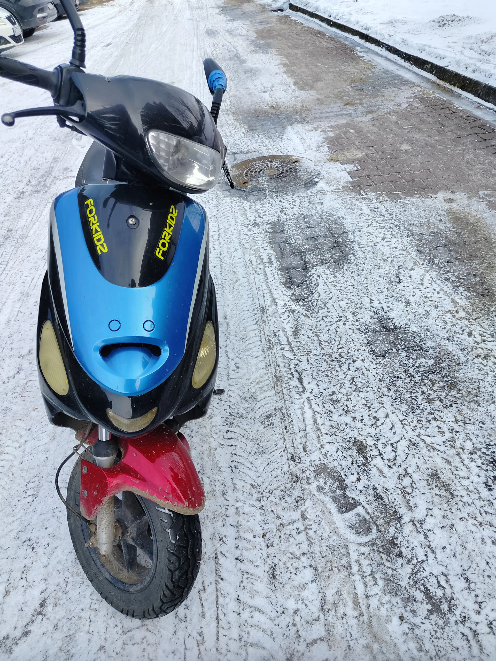 Niebiesko-czarny skuter z napisem FORKIDZ w kolorze żółtym na przedniej części, czerwonym zderzakiem i reflektorem. Skuter stoi na śnieżnej nawierzchni z widocznymi odciskami opon. W tle chodnik z częściowo oczyszczoną nawierzchnią, pokryty śniegiem, oraz kanałowa pokrywa. Po lewej stronie widoczne są fragmenty zaparkowanych samochodów.